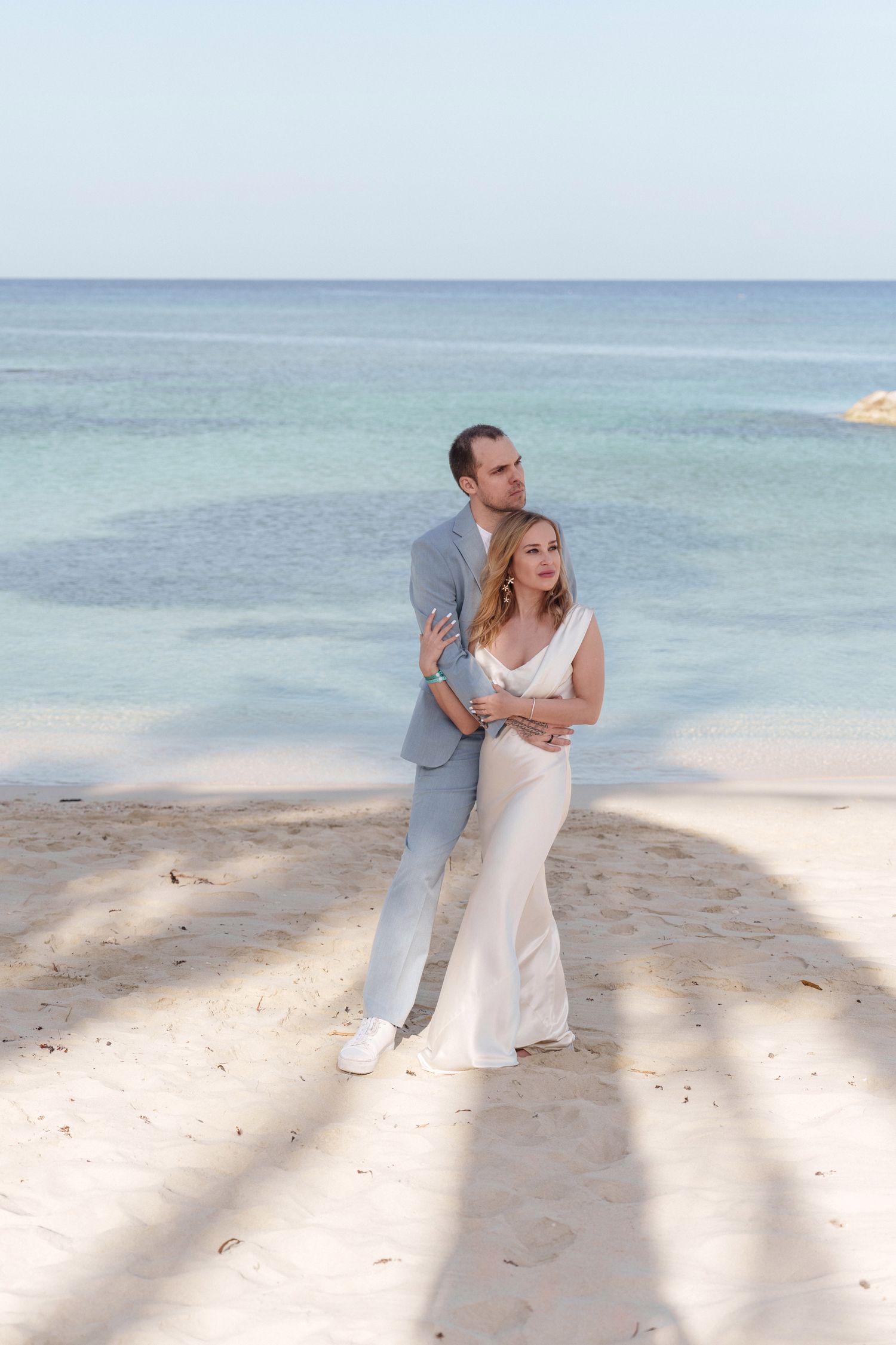 A couple in coordinated gray and white outfits embrace on a sun-dappled tropical beach.