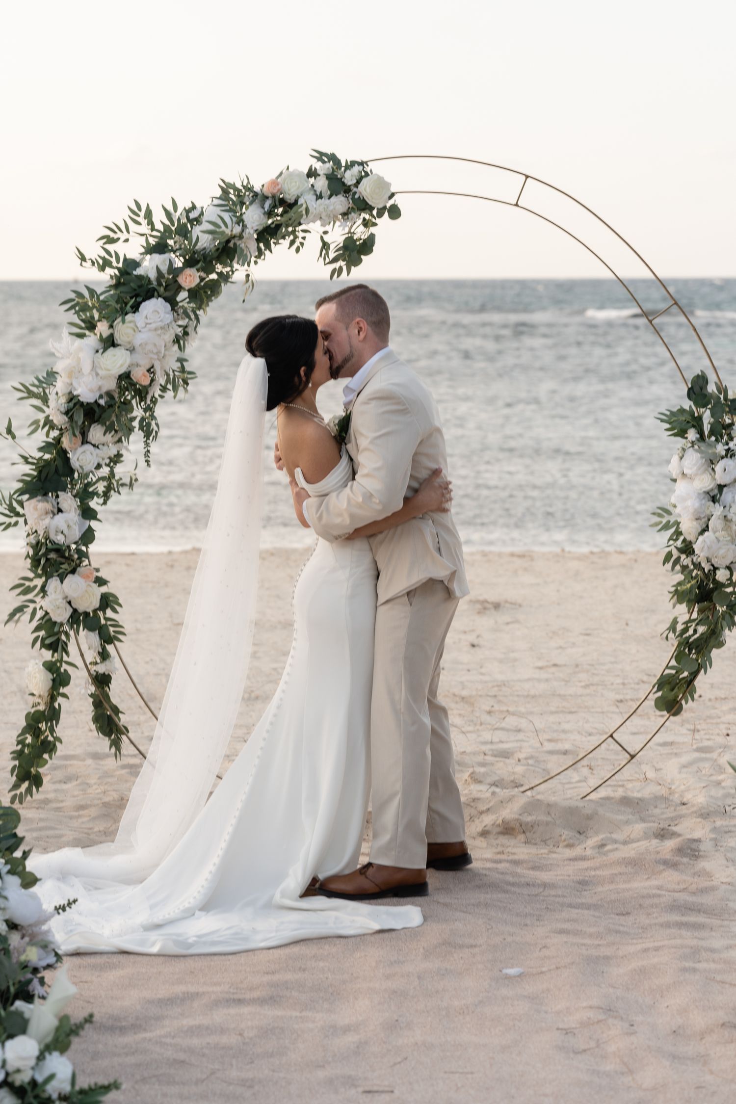 A wedding ceremony under a circular arch decorated with white flowers and greenery on the beach.