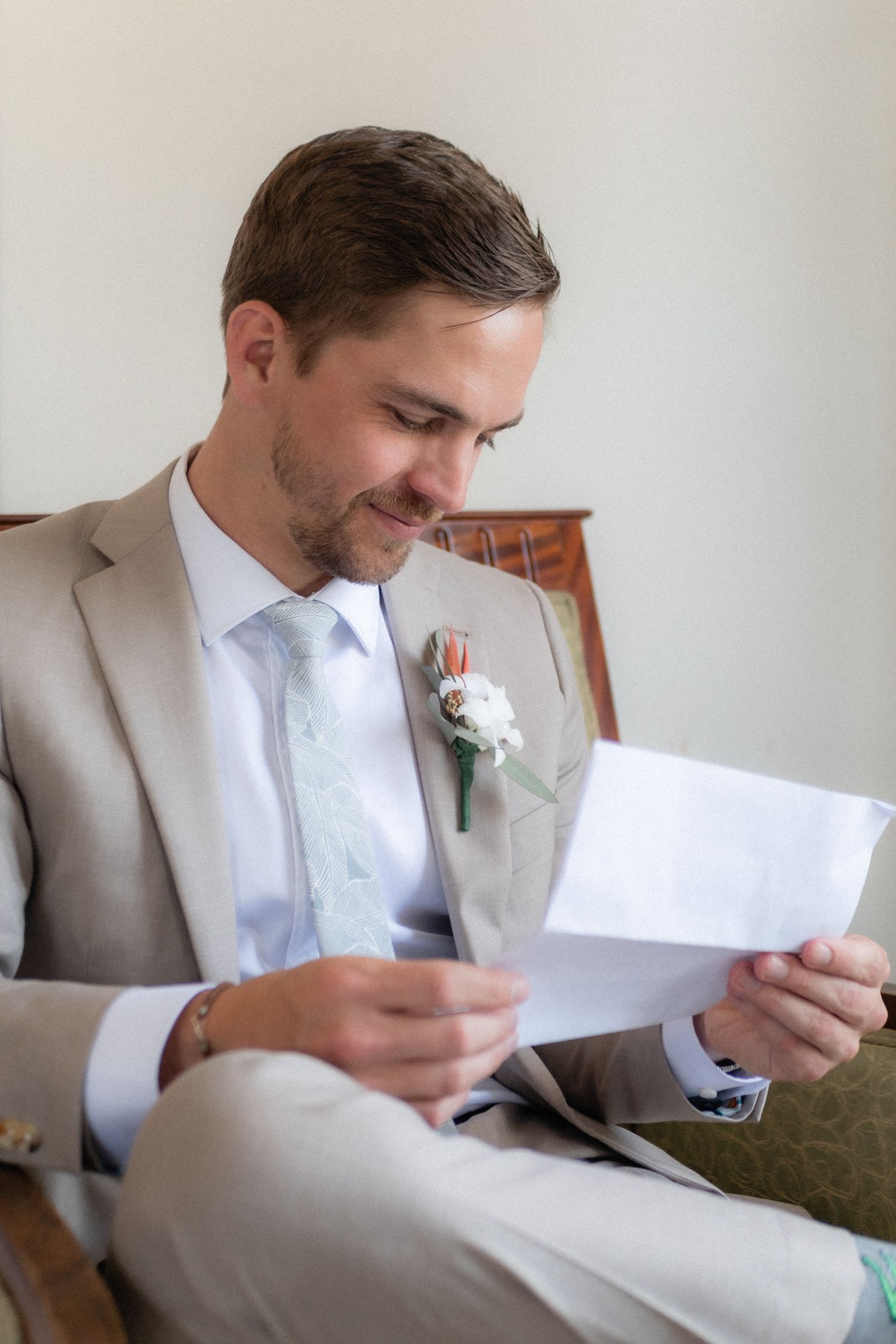 Groom in light gray suit reads letter while seated, wearing white boutonniere.