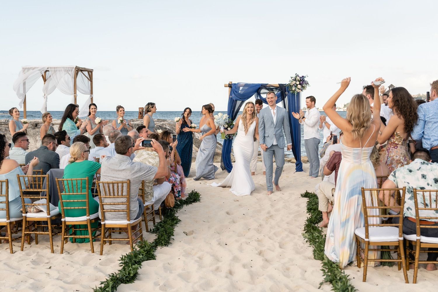A beach wedding ceremony with guests rising from chairs as couple walks down sandy aisle.