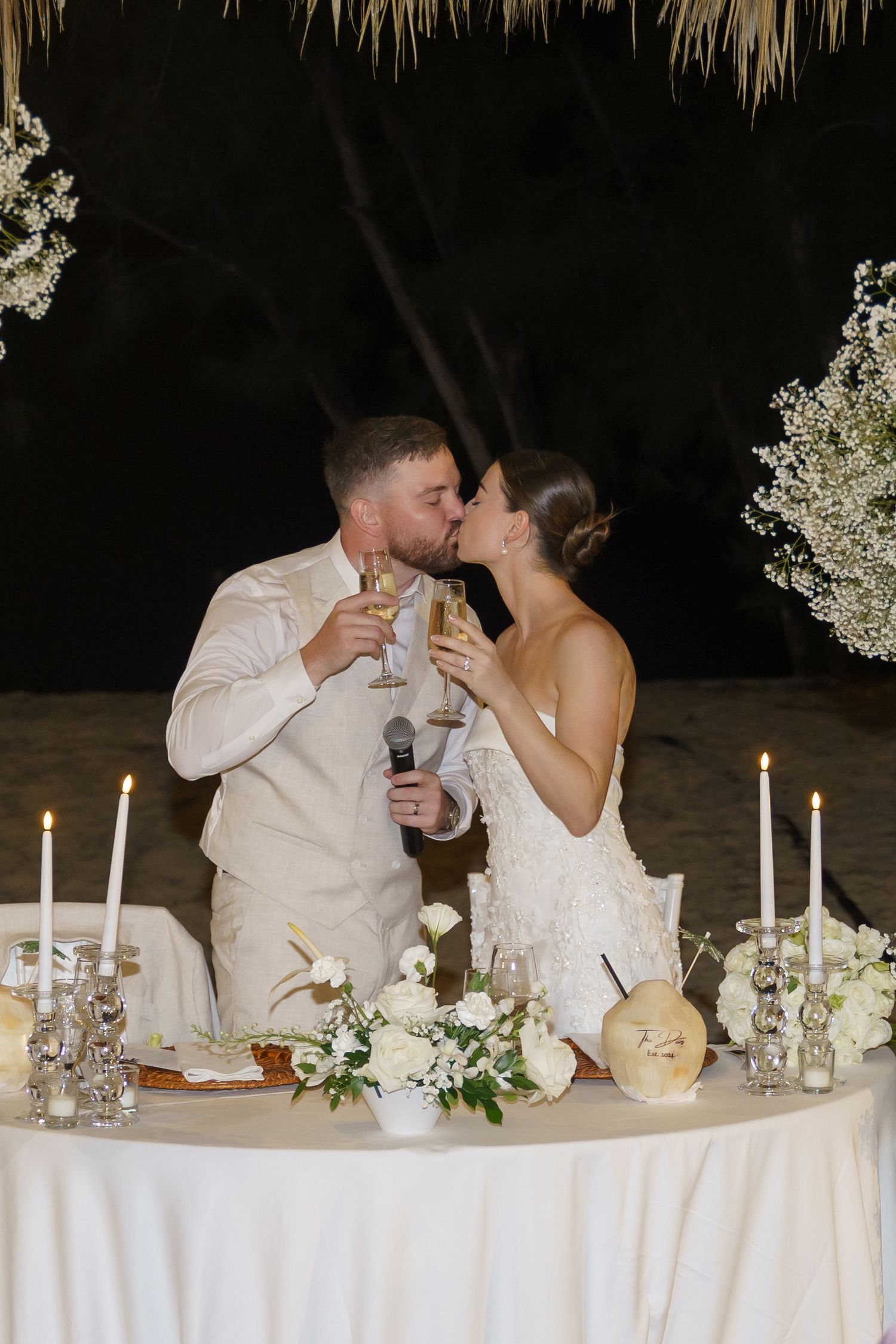 A couple kisses while toasting at their candlelit wedding reception table decorated with white flowers and linens.