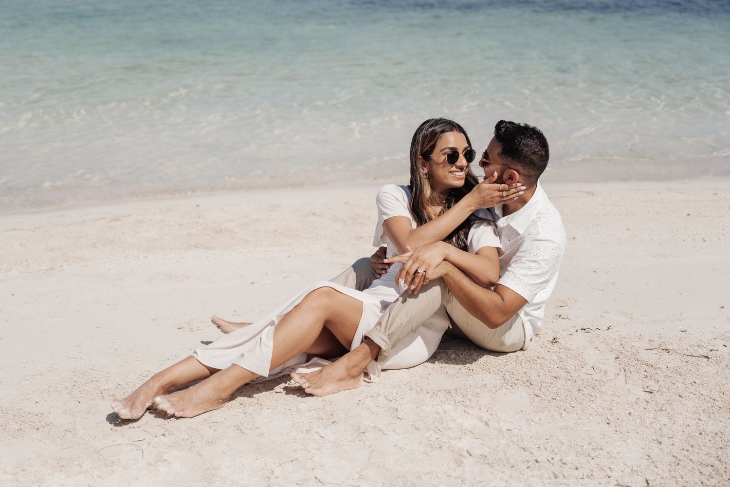 Romantic couple in white outfits lounging together on a tropical beach with crystal clear waters.
