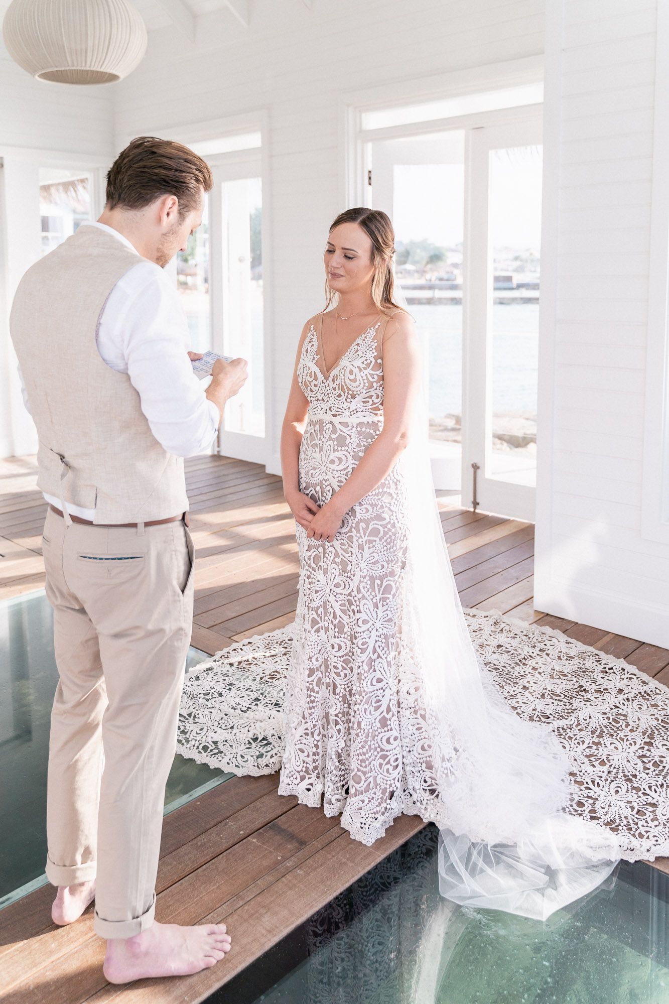 A couple exchanges vows on a glass walkway in an elegant white oceanfront venue.