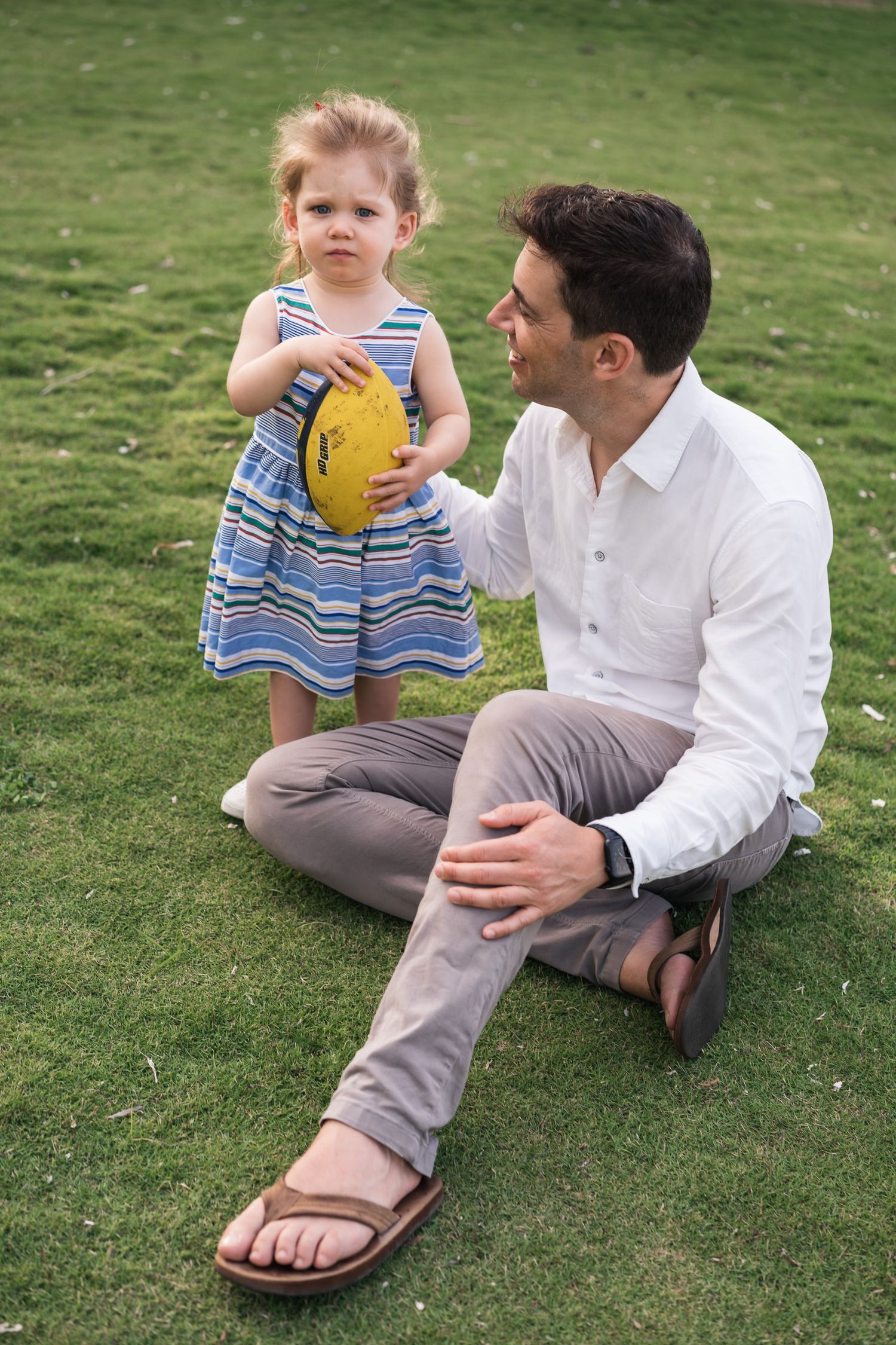Adult and child sitting on grass having a conversation while holding a yellow ball.