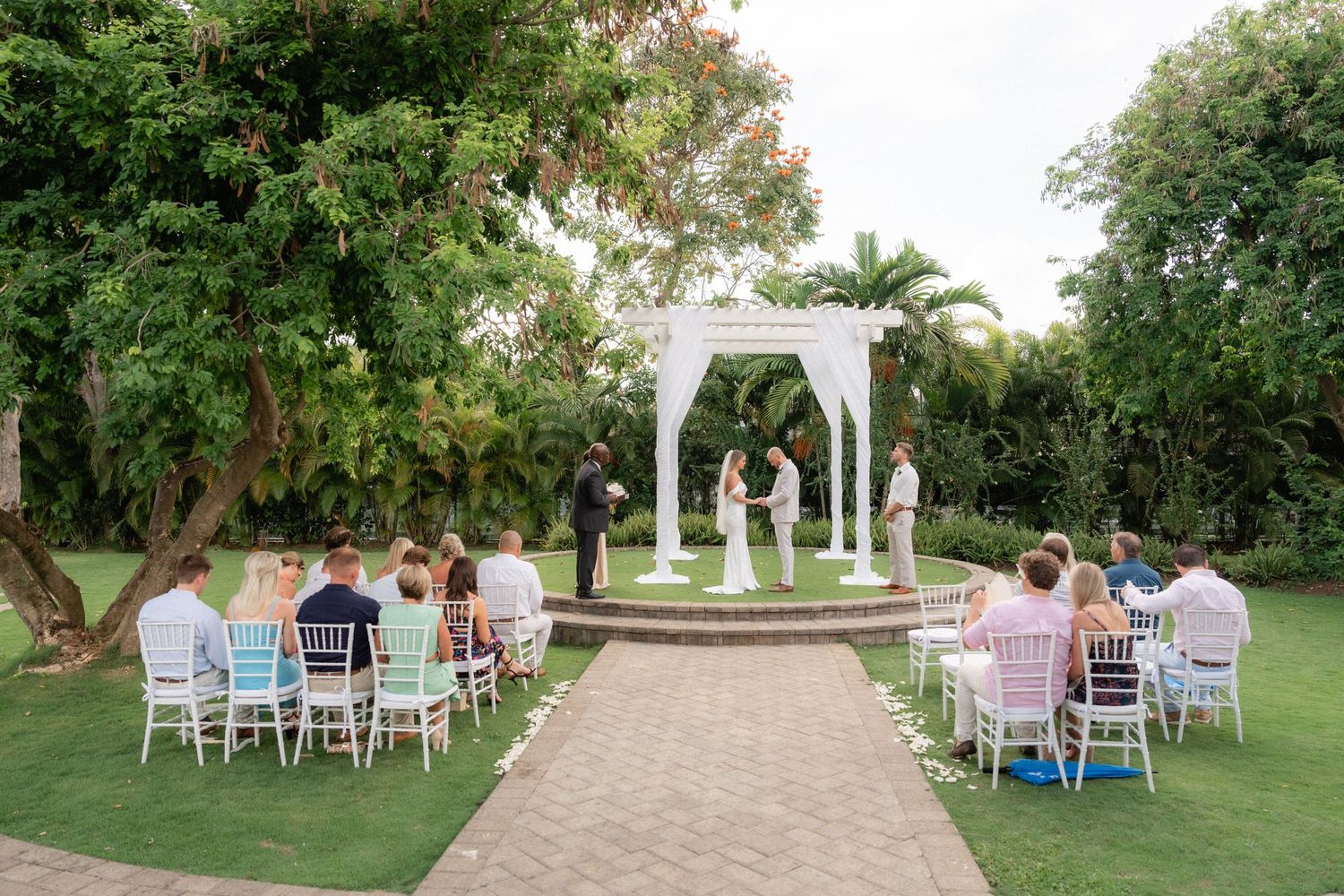 Intimate outdoor garden wedding ceremony with white gazebo and guests seated in white chairs.
