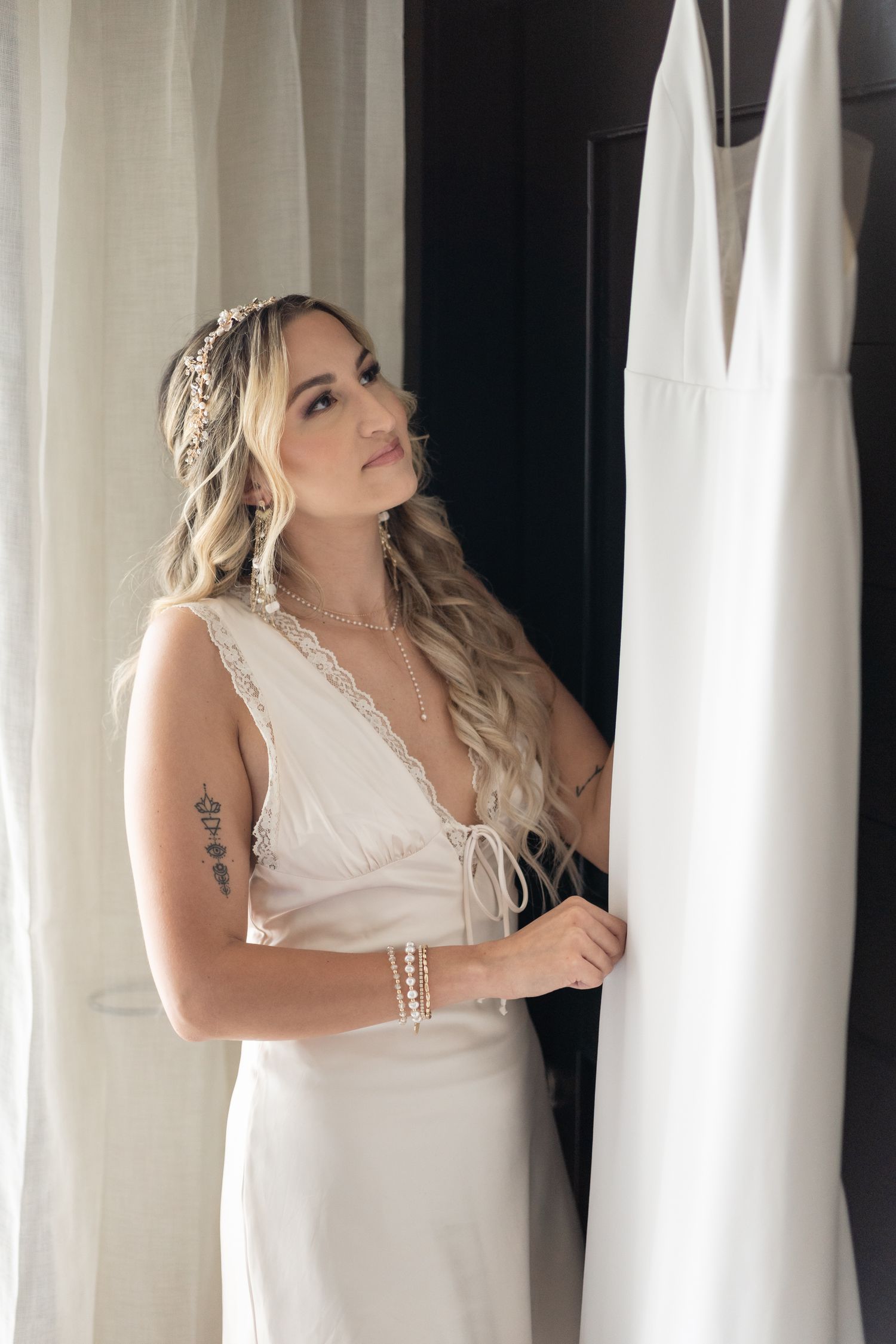 Bride in white dress with flowing blonde hair and delicate headpiece standing near curtains.
