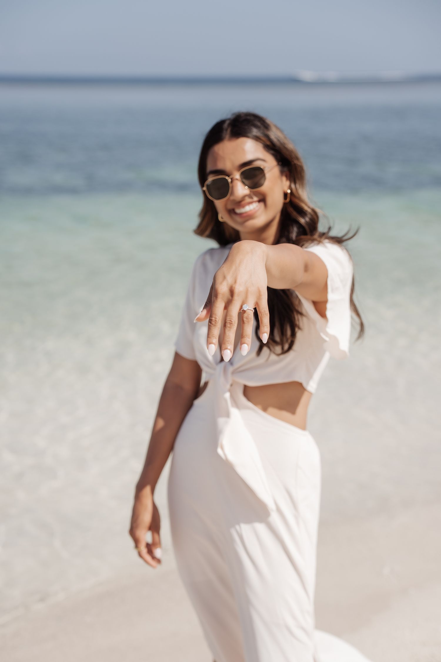 Woman in white dress shows engagement ring on tropical beach.