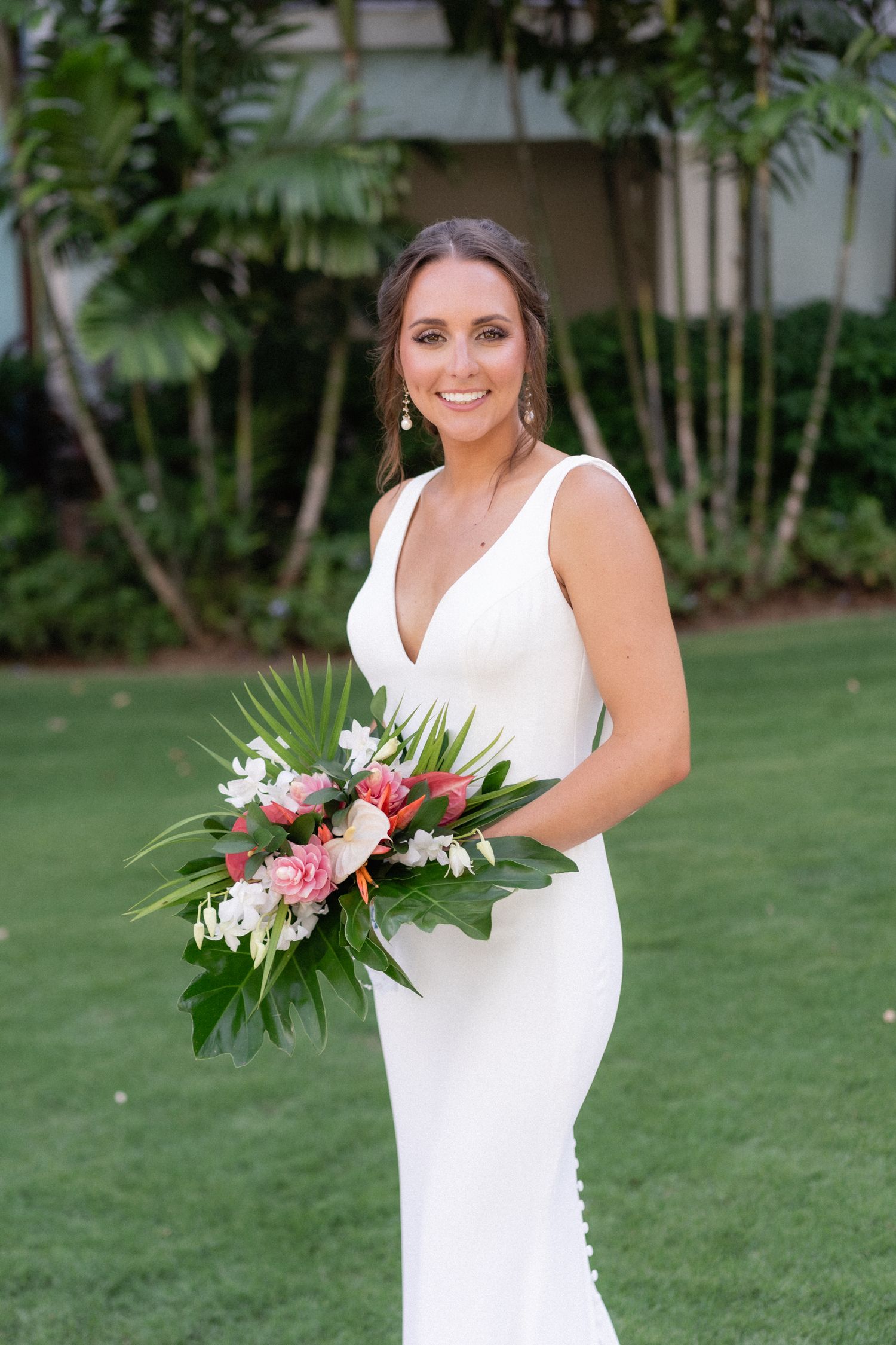 Elegant bride in fitted white v-neck dress holds tropical bouquet with pink flowers against garden backdrop.