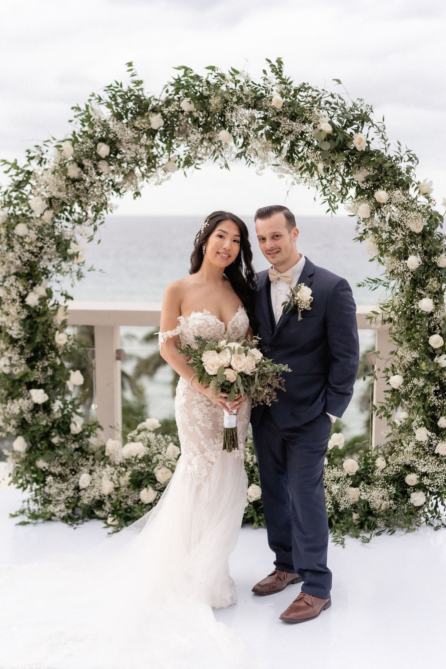 A newlywed couple poses under a lush white floral arch with an ocean view.