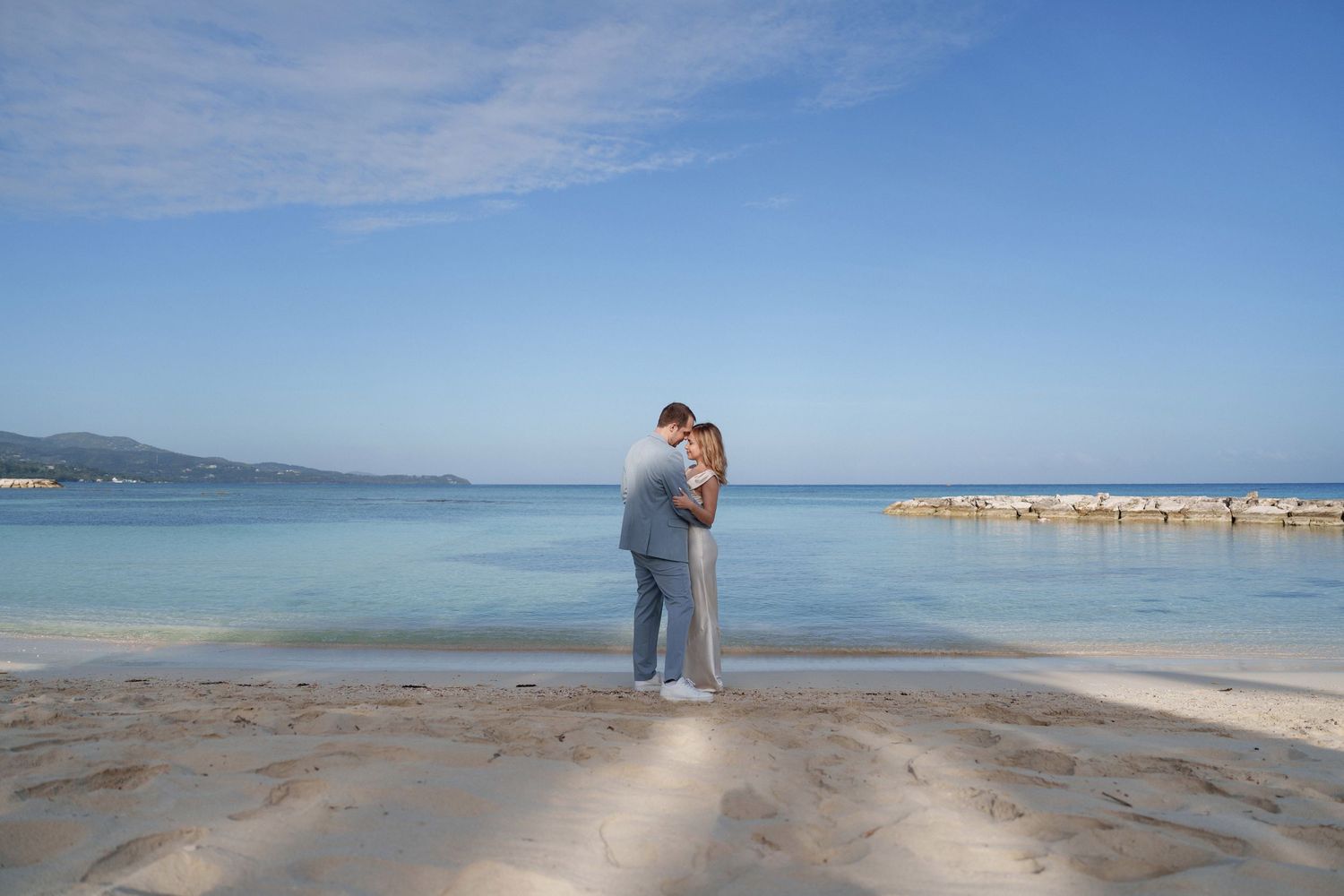 Two people in matching gray outfits share a tender moment on a tropical beach with mountains in the distance.