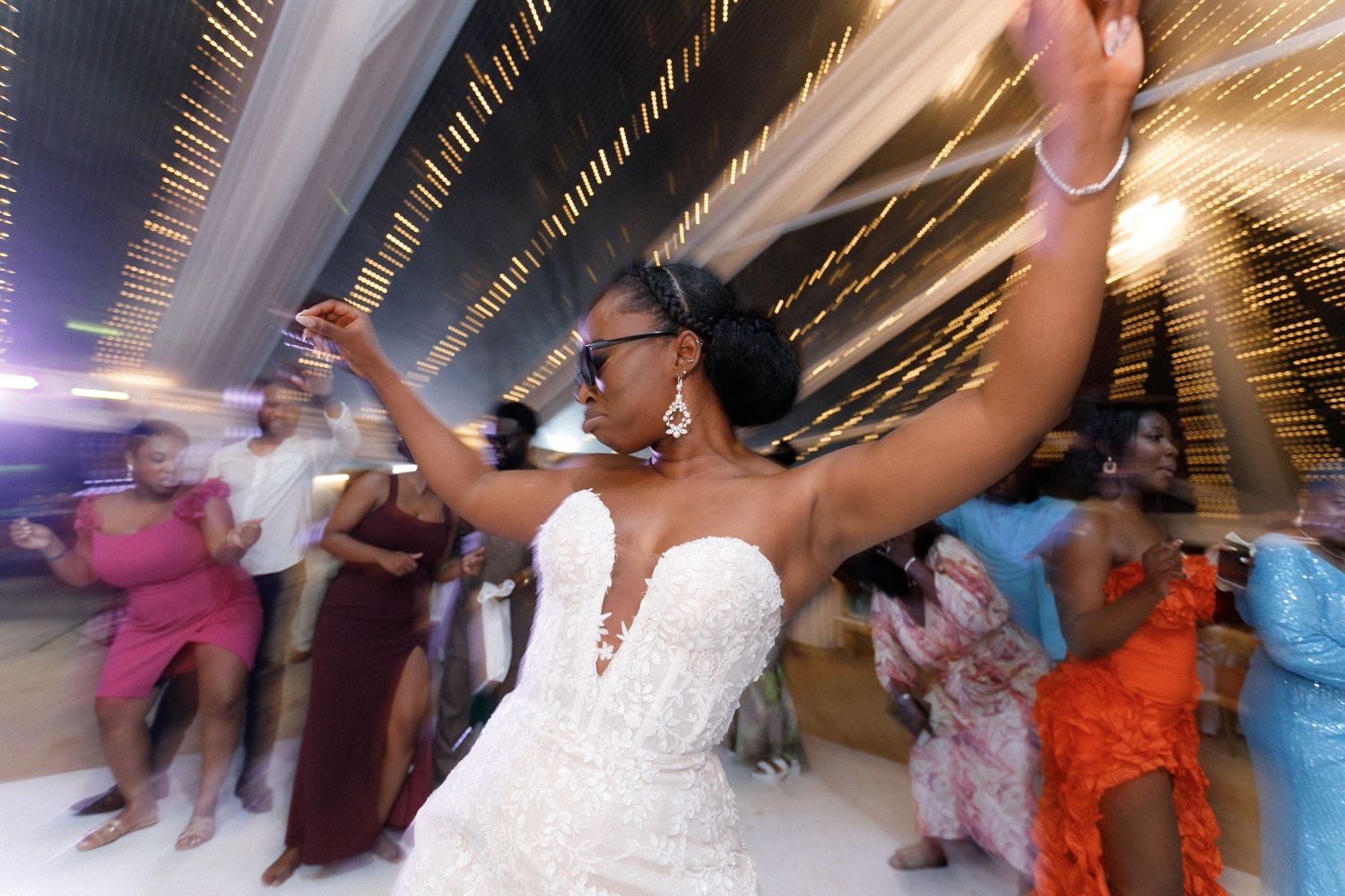 Motion-blurred shot of wedding guests dancing energetically under decorative string lights.