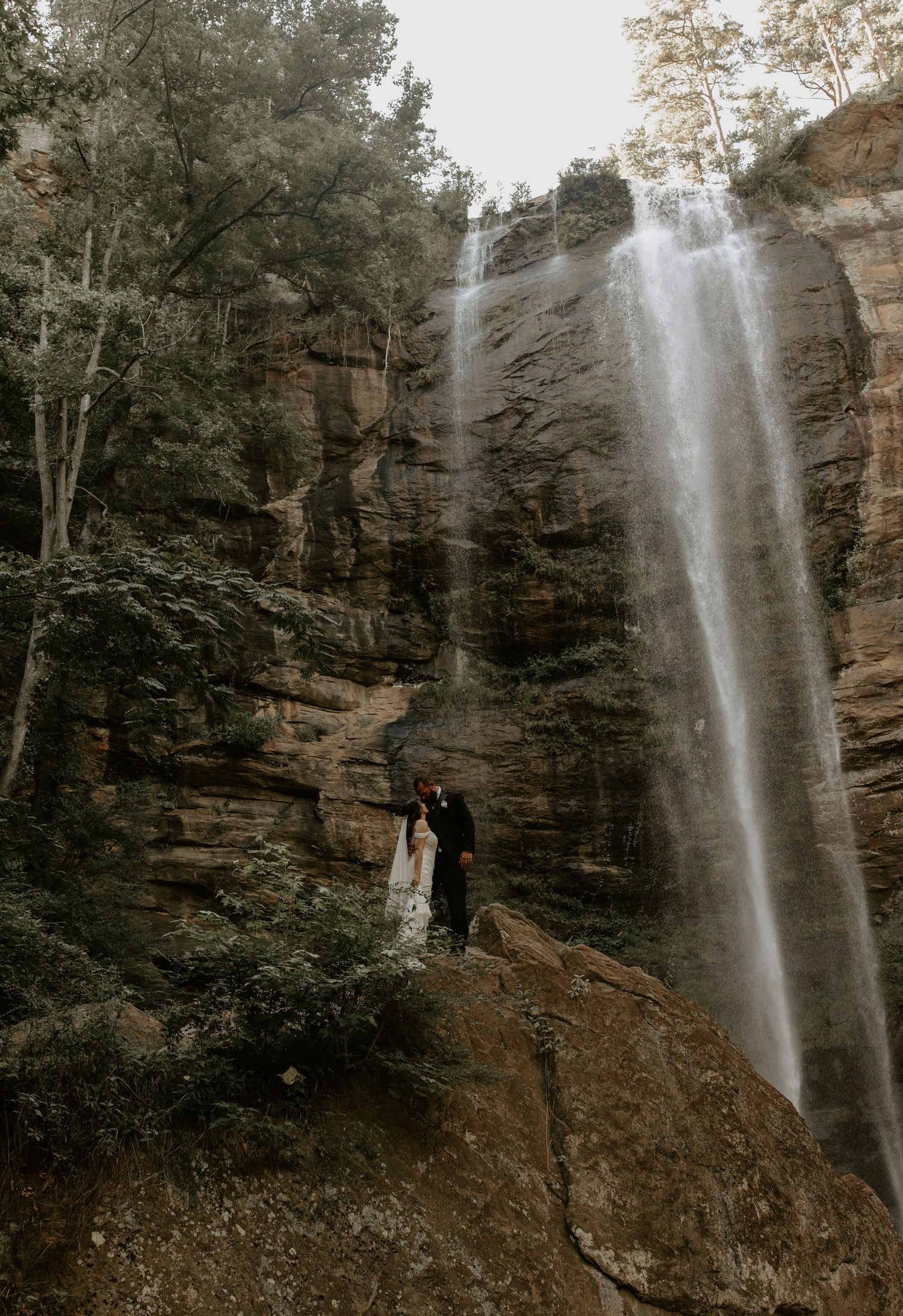 A romantic couple stands together beneath a majestic waterfall cascading down tall rocky cliffs in a misty forest setting.