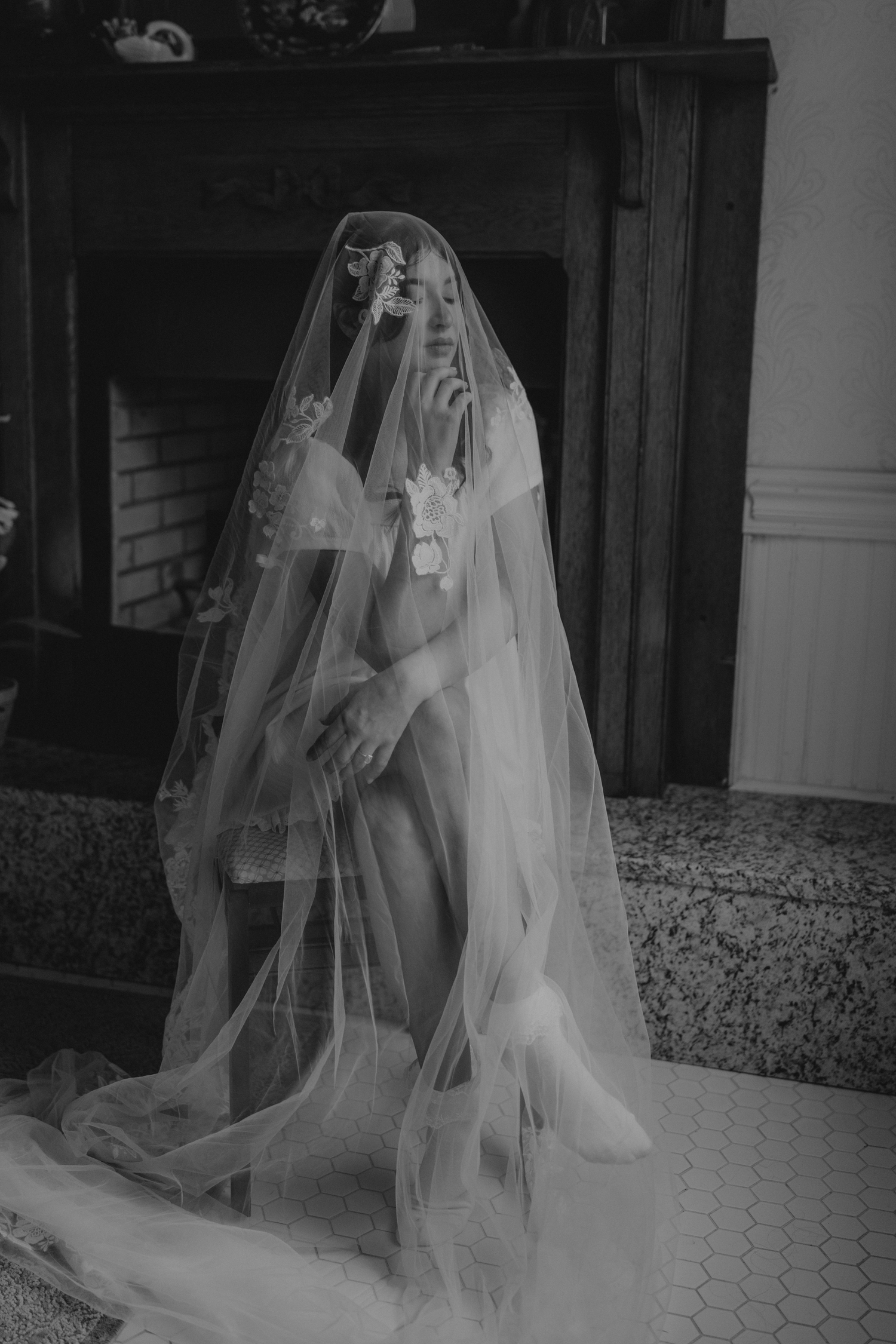 Black and white photo of a bride wearing a flowing veil and floral crown, seated near a fireplace.