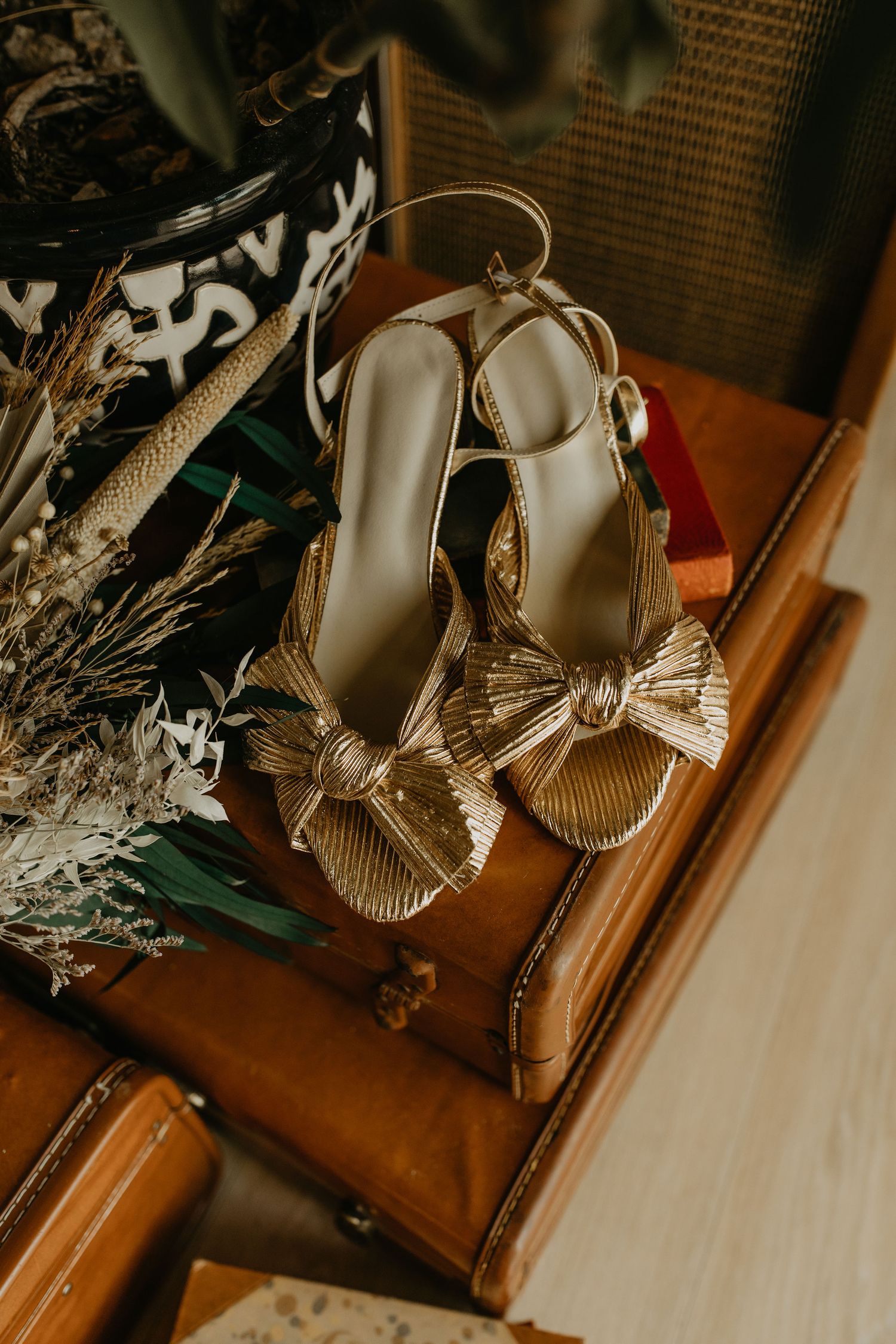Elegant gold metallic sandals with bow detail displayed on vintage brown leather case with dried floral decorations.