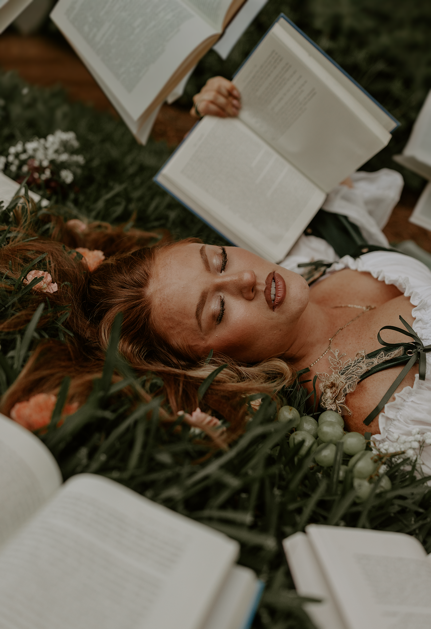 Woman with red hair lying in grass surrounded by open books, wearing a white dress with floral accessories.
