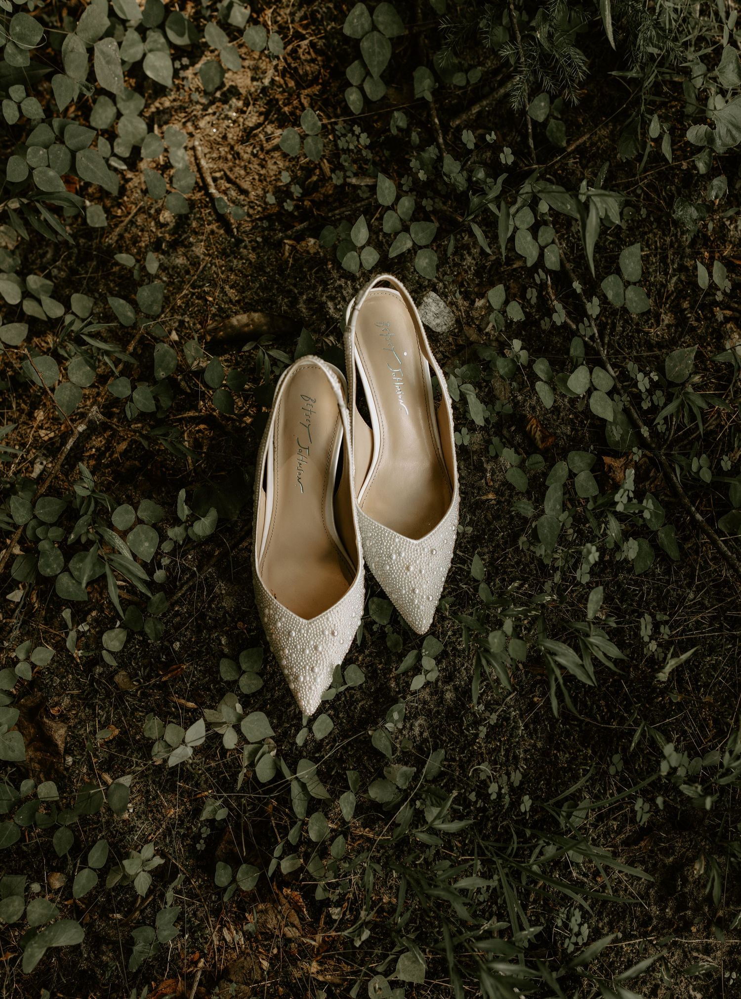 Elegant white bridal shoes with delicate pearl embellishments rest on dark green foliage backdrop.