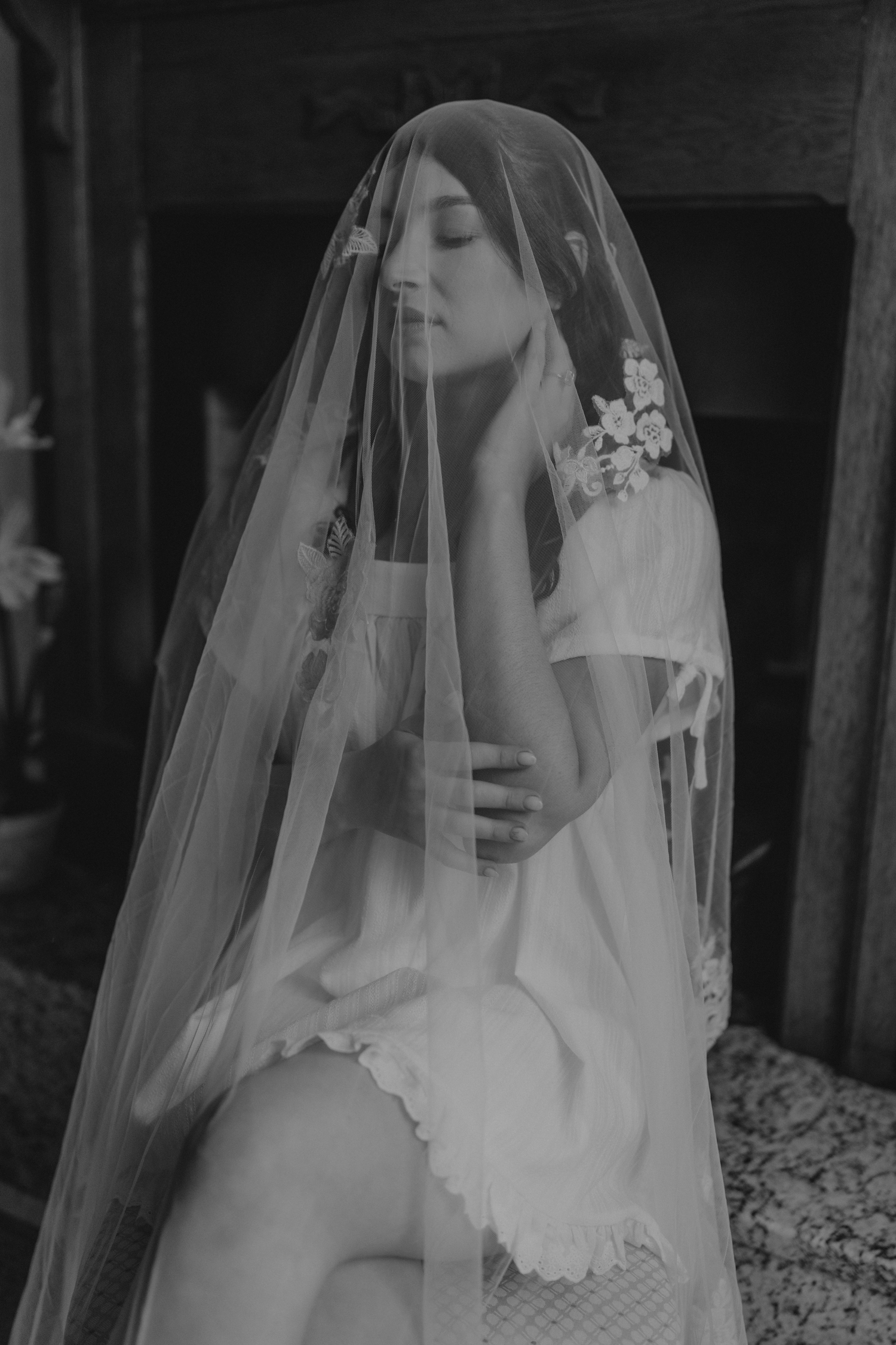 Black and white photo of a bride in white dress and veil with floral accents, sitting gracefully with eyes closed.