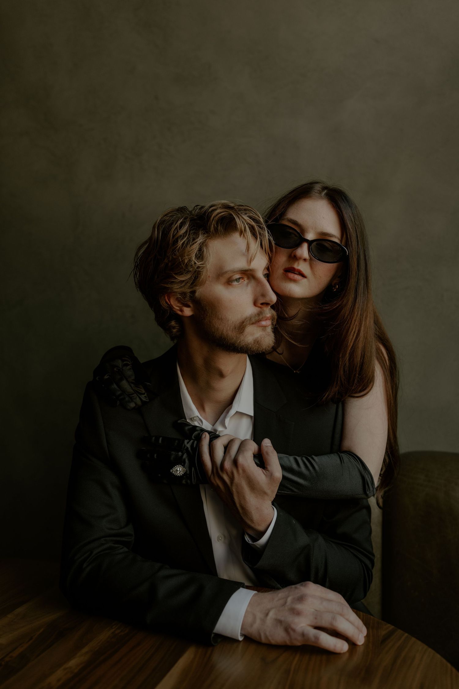 A stylish couple poses intimately at a wooden table, man in dark suit, woman in sunglasses embracing him from behind.