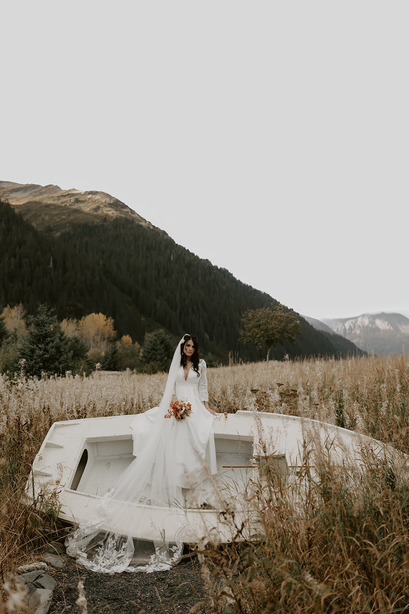 Bride in white dress standing on rustic boat in mountain field landscape.