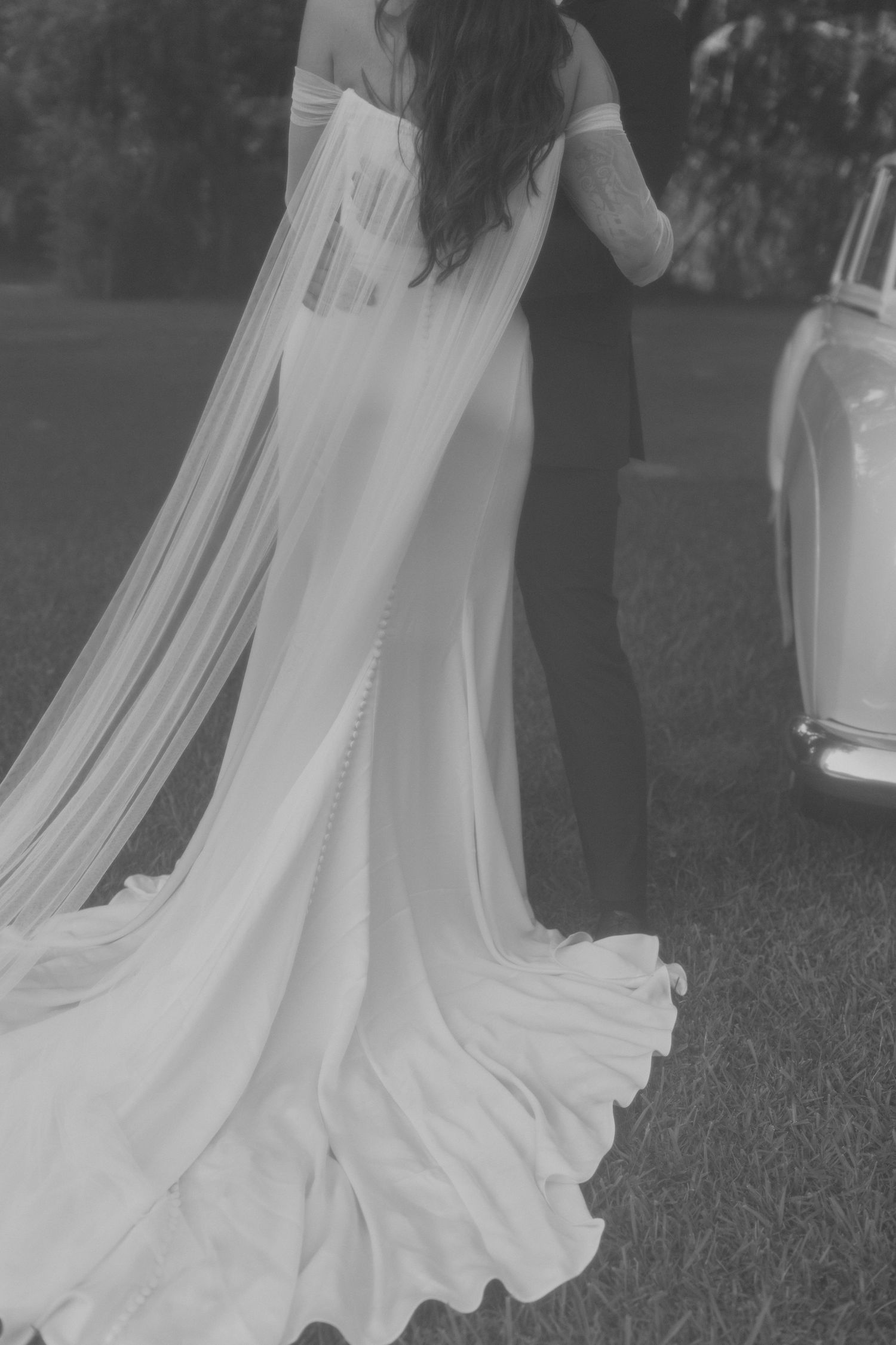 Close-up black and white shot of flowing wedding dress train against vintage car.