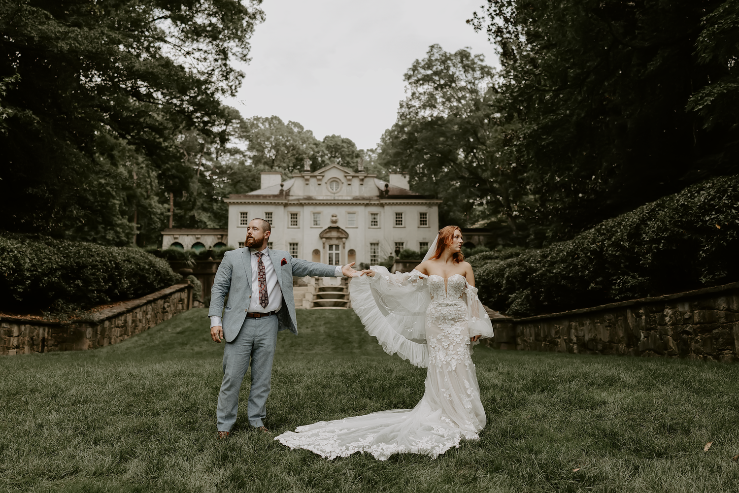 A couple poses outdoors in formal wedding attire against a backdrop of lush greenery and a white mansion.