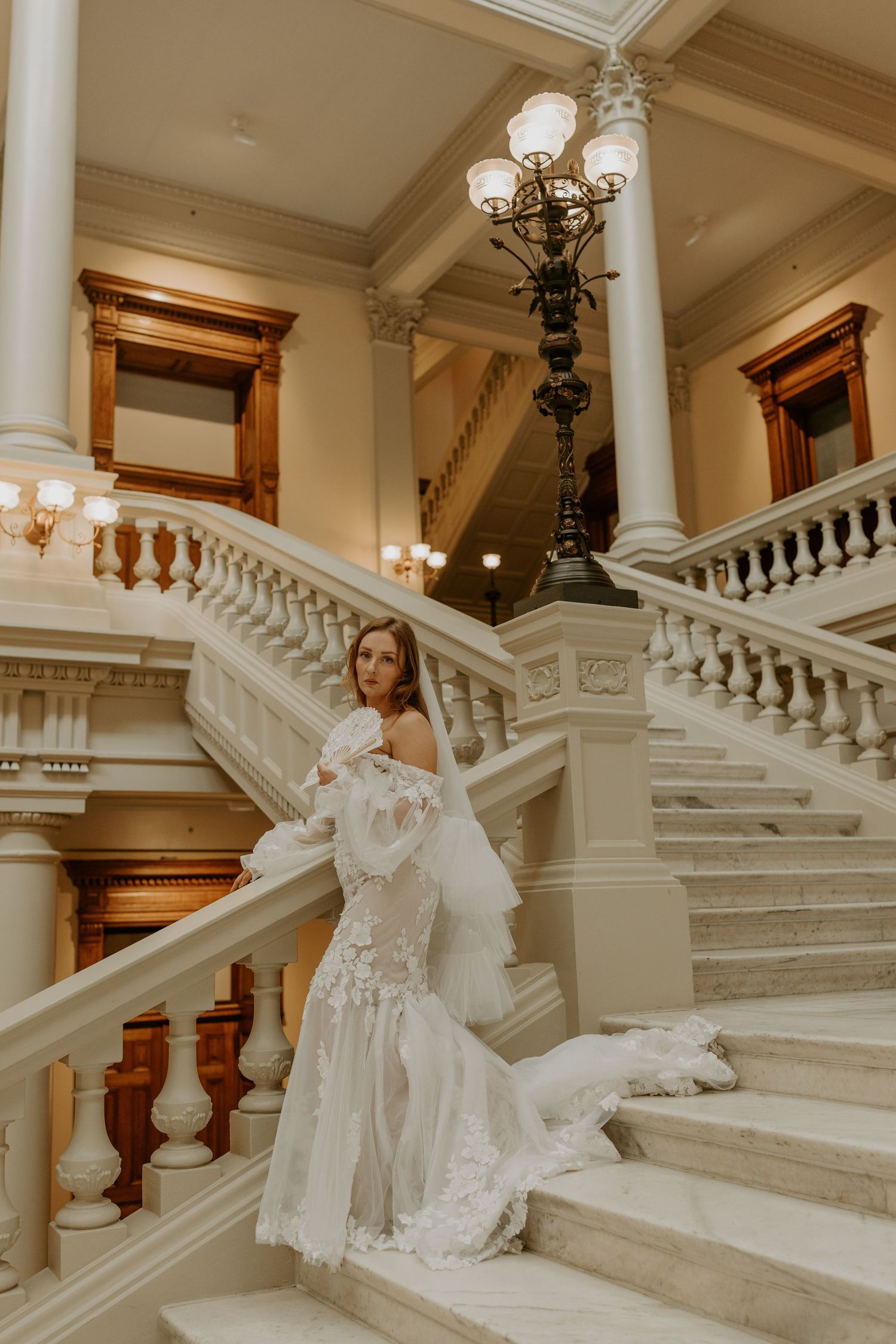 A bride in a white lace gown poses elegantly on ornate marble stairs inside a grand historic building with classic architecture.