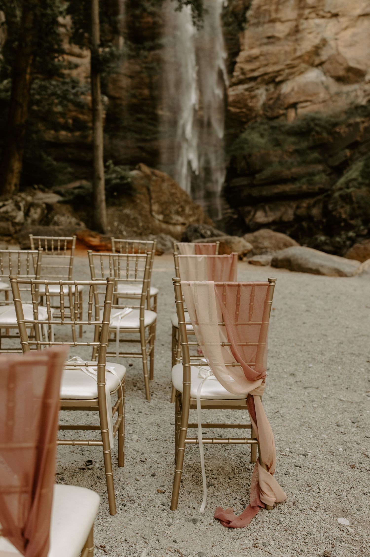 Gold chiavari chairs with blush drapes set up for an outdoor wedding ceremony against dramatic rock cliff backdrop.