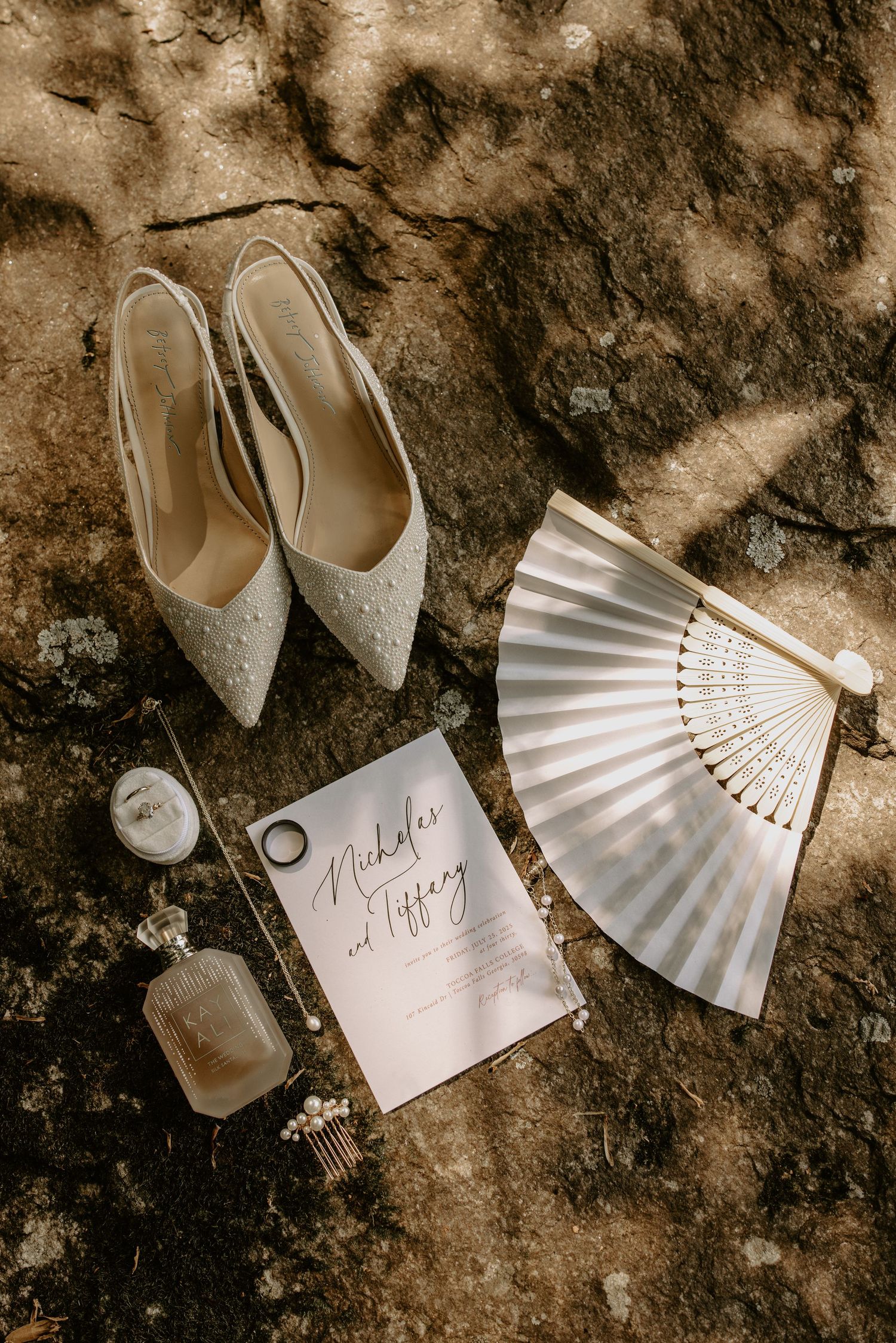 Stylish wedding accessories including elegant white heels, paper fan, and handwritten note arranged on textured stone background.