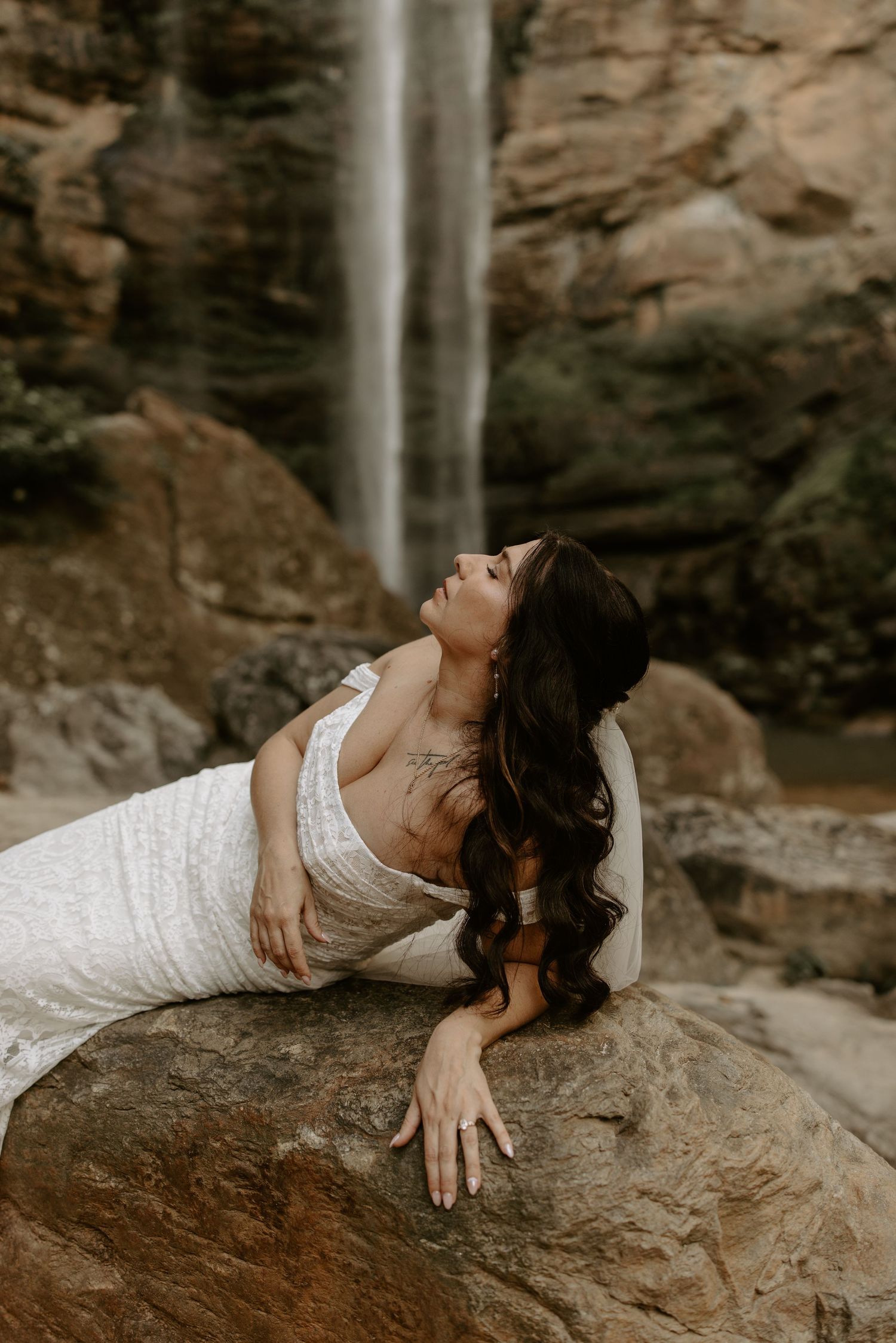 A figure in a white dress reclines gracefully on rocky terrain near a waterfall in a dramatic natural setting.