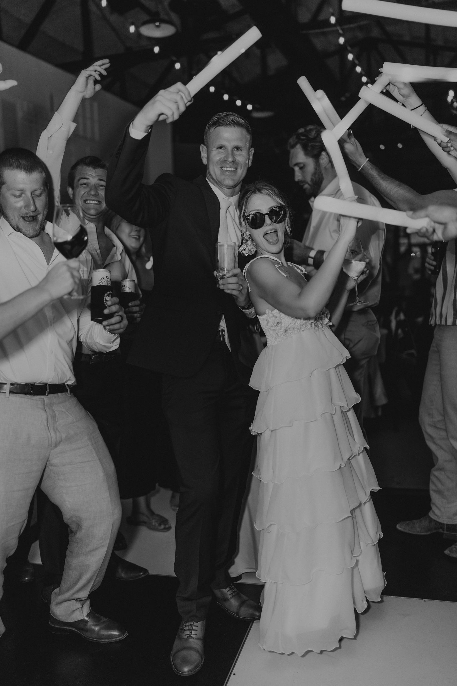 Wedding guests dance and celebrate with foam glow sticks while bride wears tiered white dress and sunglasses at reception.