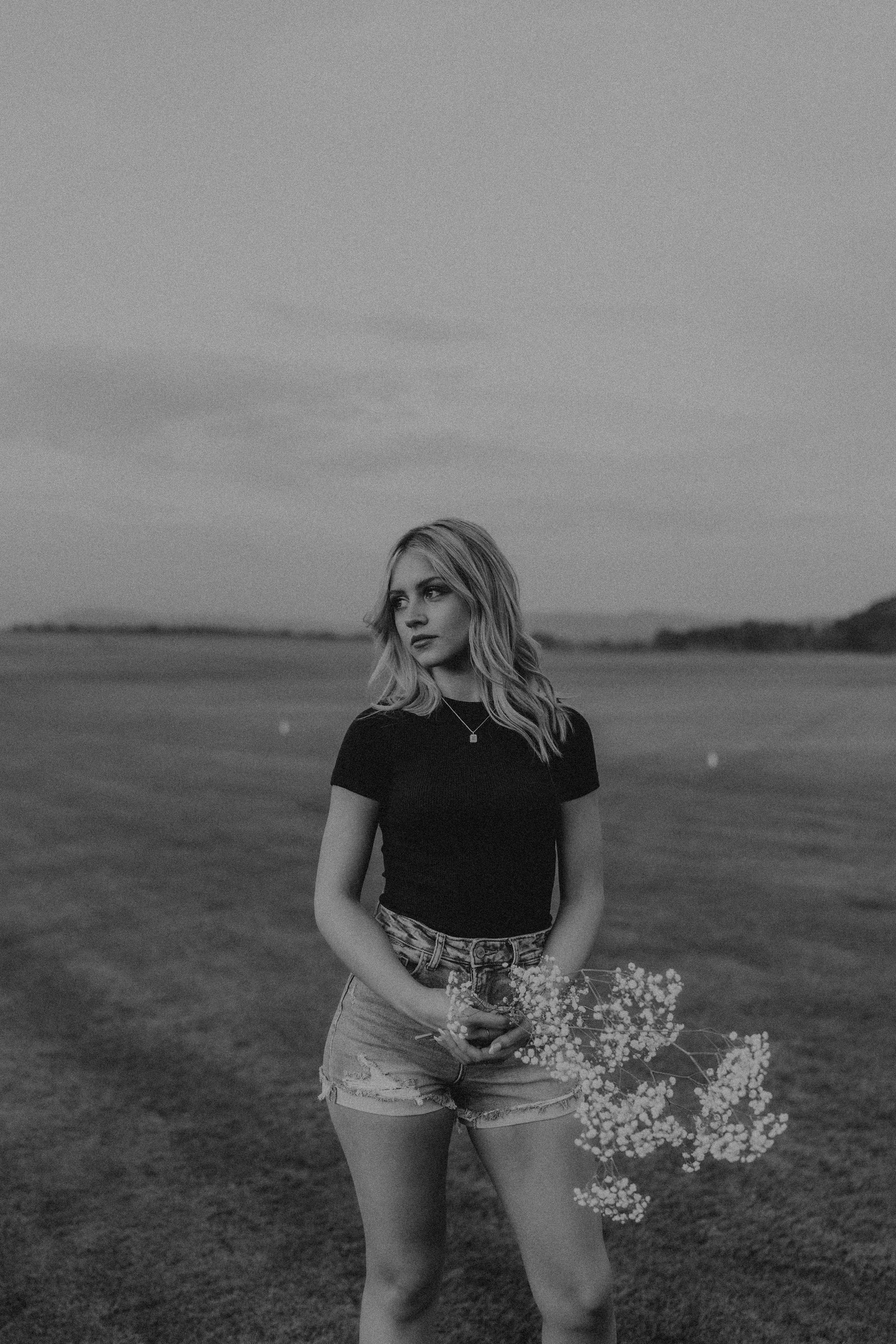 A black and white photo of someone standing in denim shorts and black top holding small wildflowers in an open field.