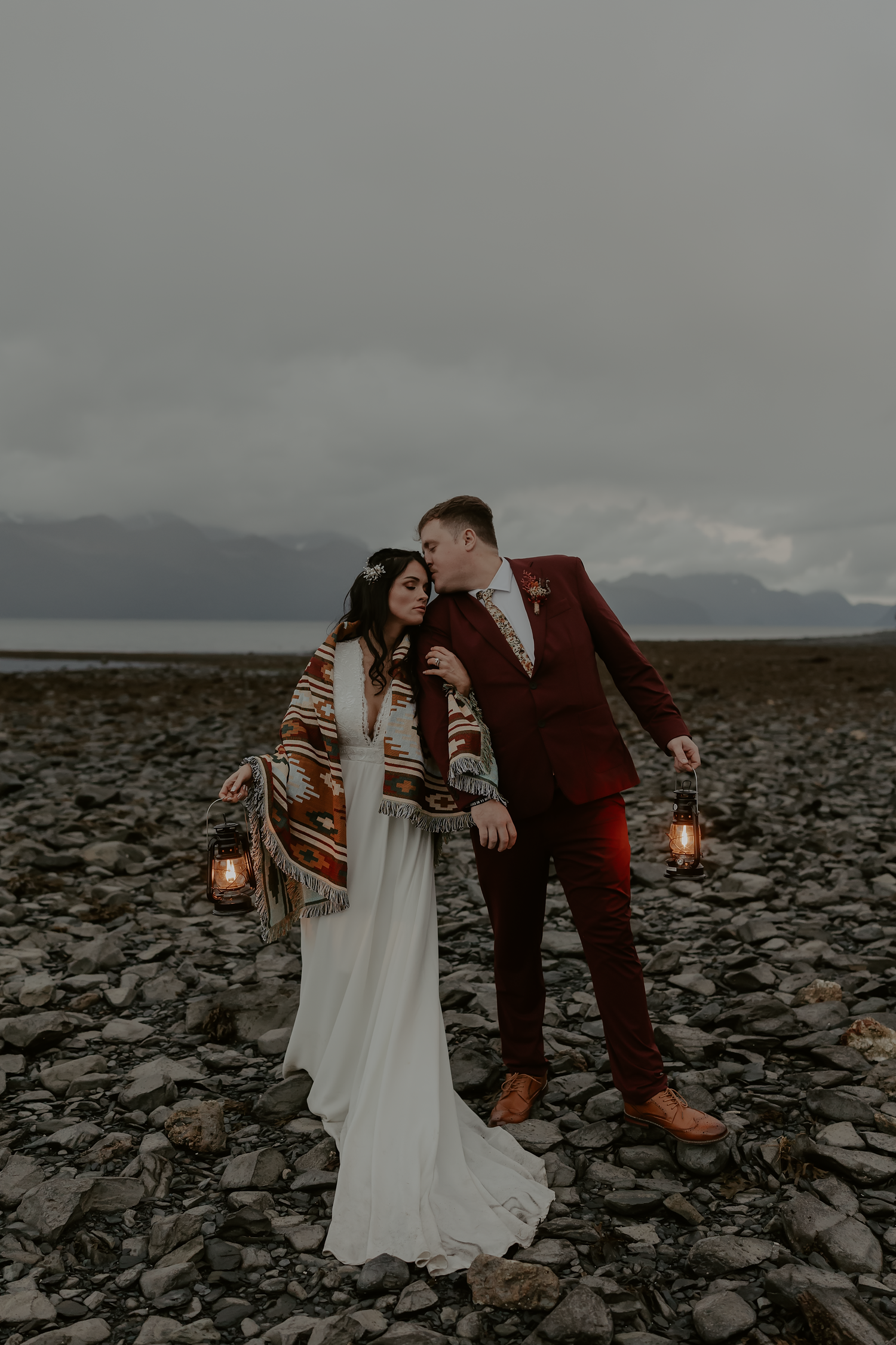 Couple in wedding attire share intimate moments on rocky terrain at dusk while holding vintage lanterns.