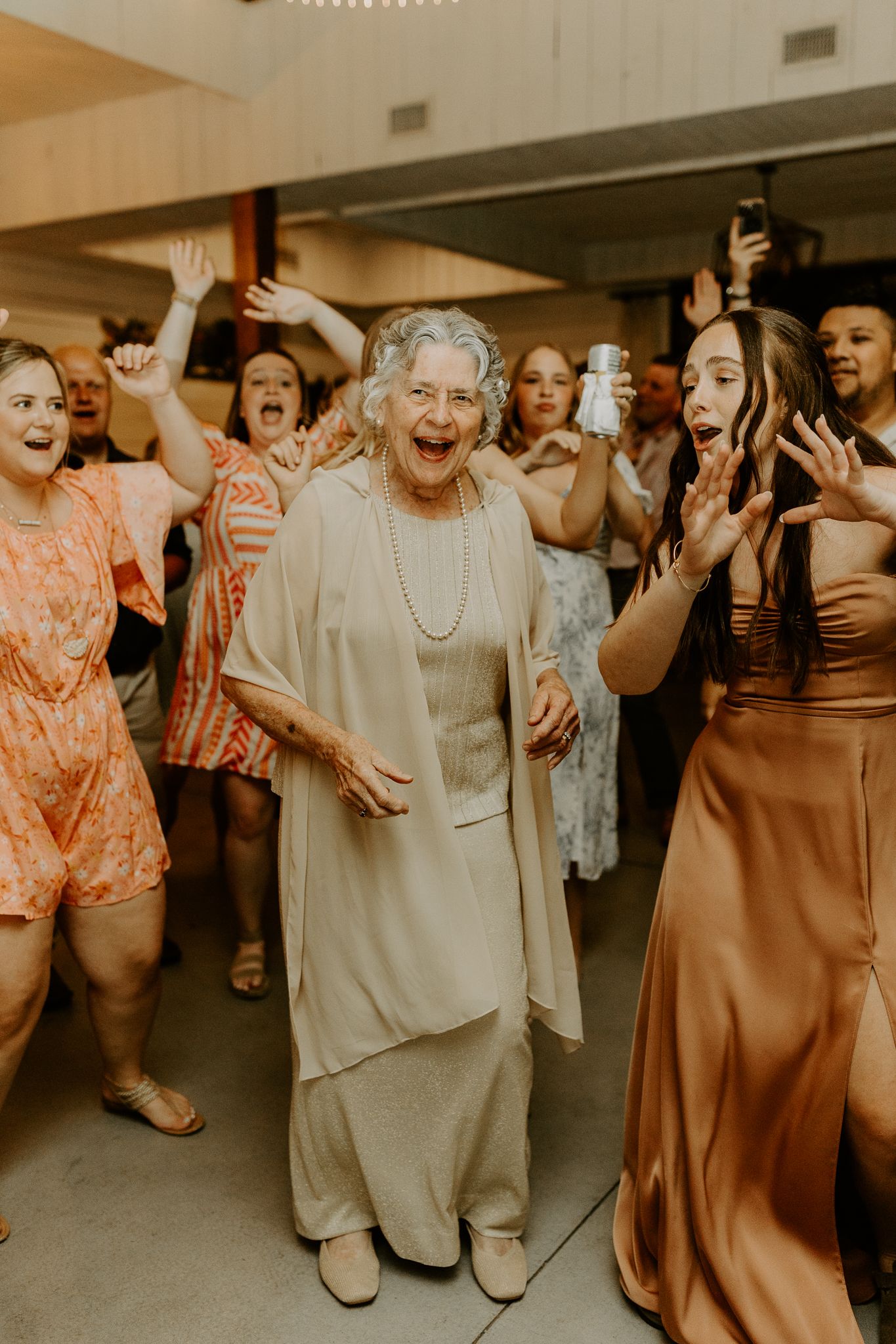 A joyful group of wedding guests dancing energetically at a reception with happy expressions and raised arms.