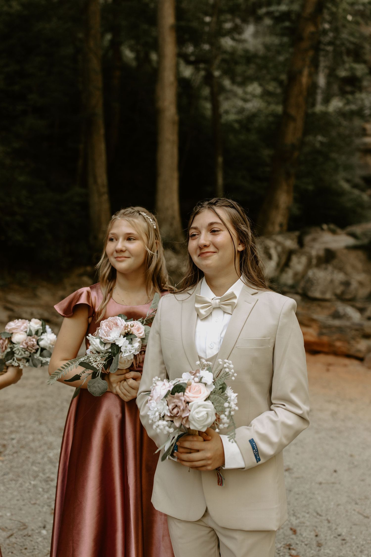 Two people in coordinating wedding attire hold bouquets while standing together in a forest setting.