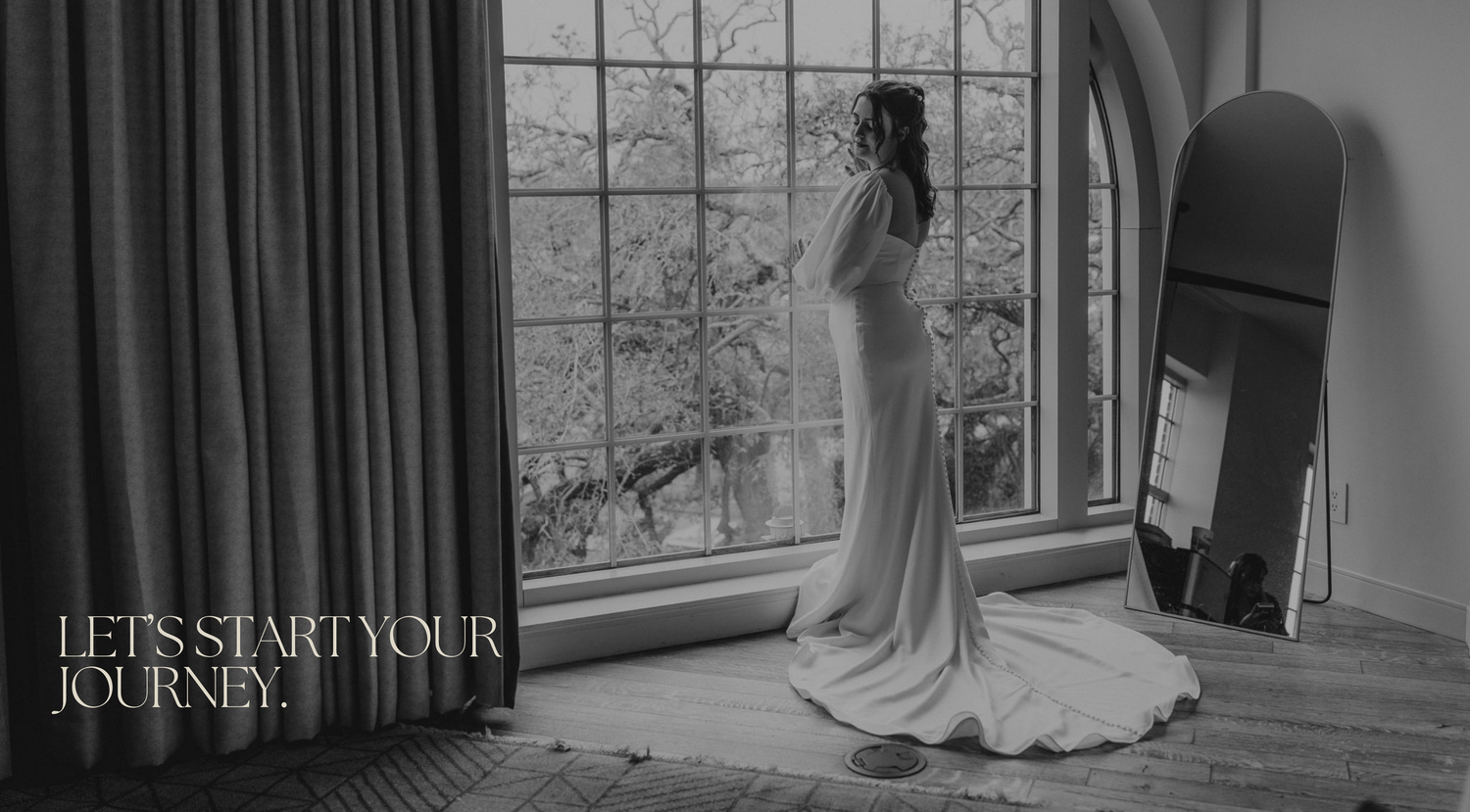 Bride in elegant white gown standing by large window in black and white wedding photo with text Let's Start Your Journey.