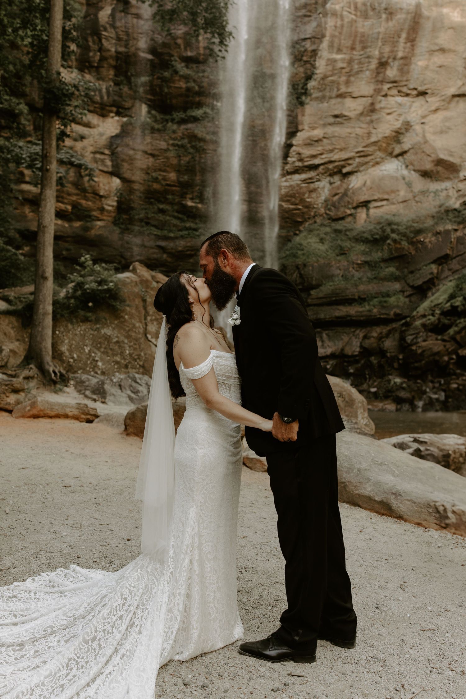 A couple in wedding attire share a tender moment in front of a majestic waterfall against rocky cliffs.
