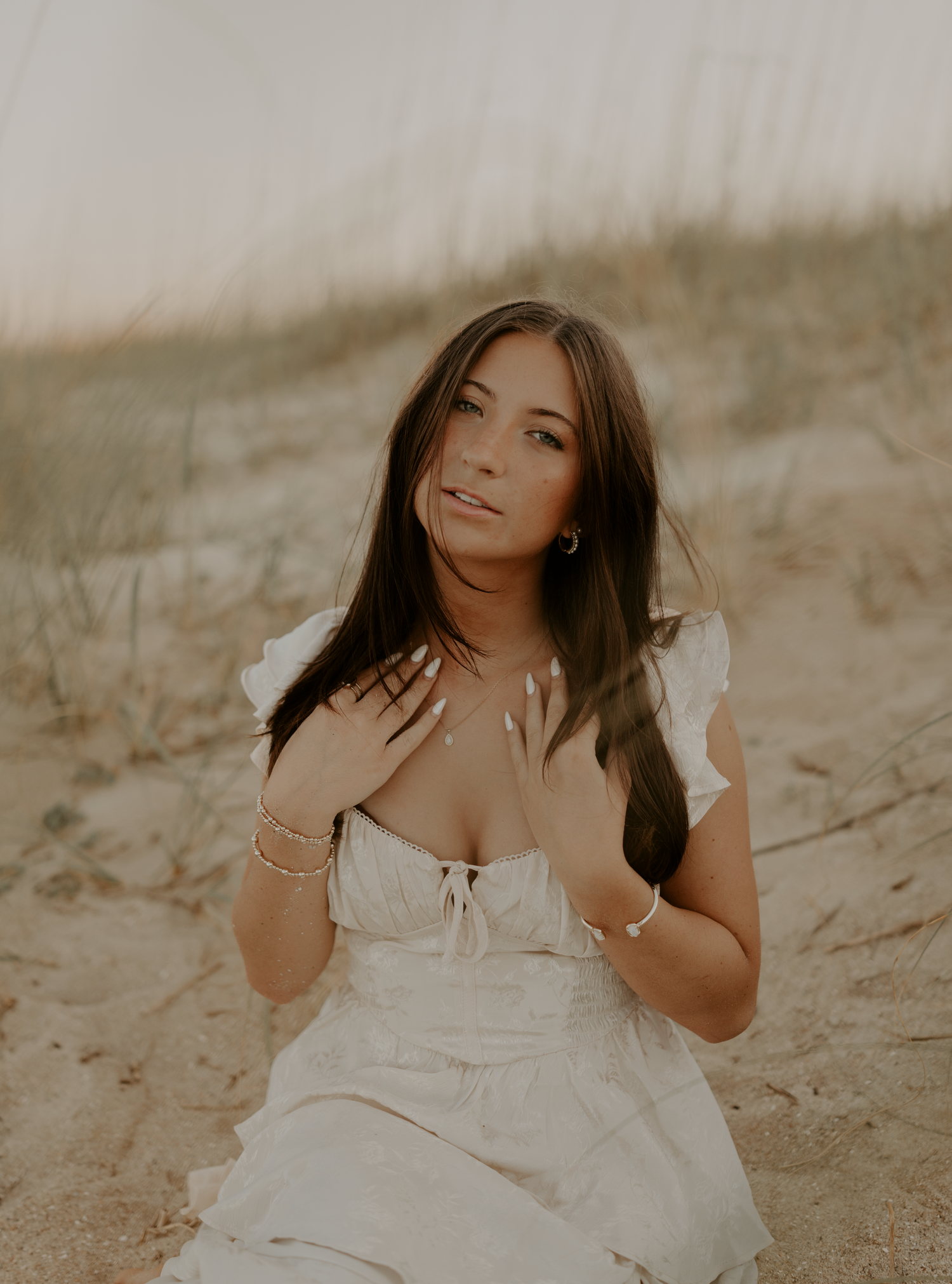 Young woman in white ruffled dress sitting on sandy beach dunes, golden hour light, long dark hair, gold jewelry.