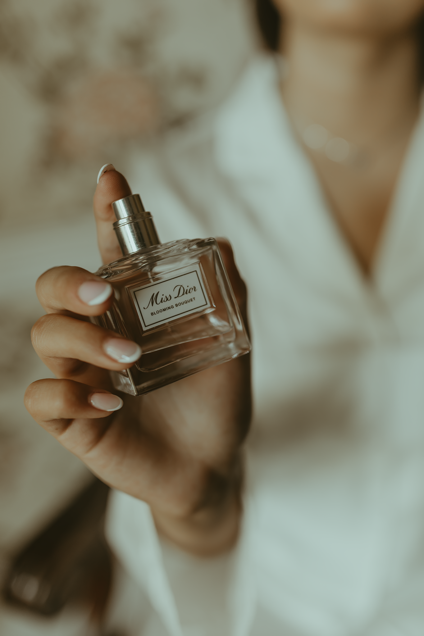 Close-up shot of a hand holding a Miss Dior perfume bottle with manicured nails in soft, dreamy lighting.