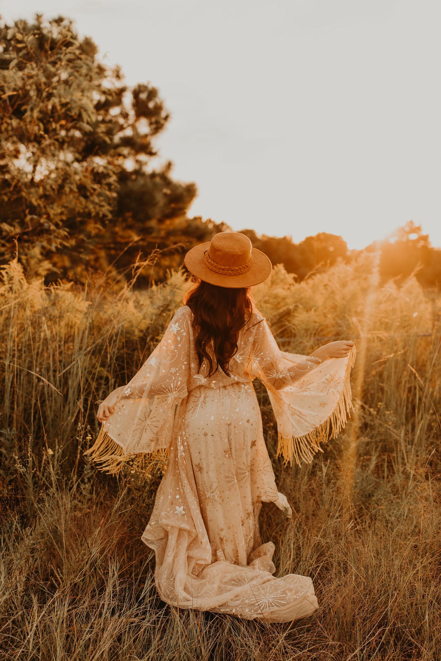 A person in a flowing white vintage dress and straw hat stands in a golden field at sunset.