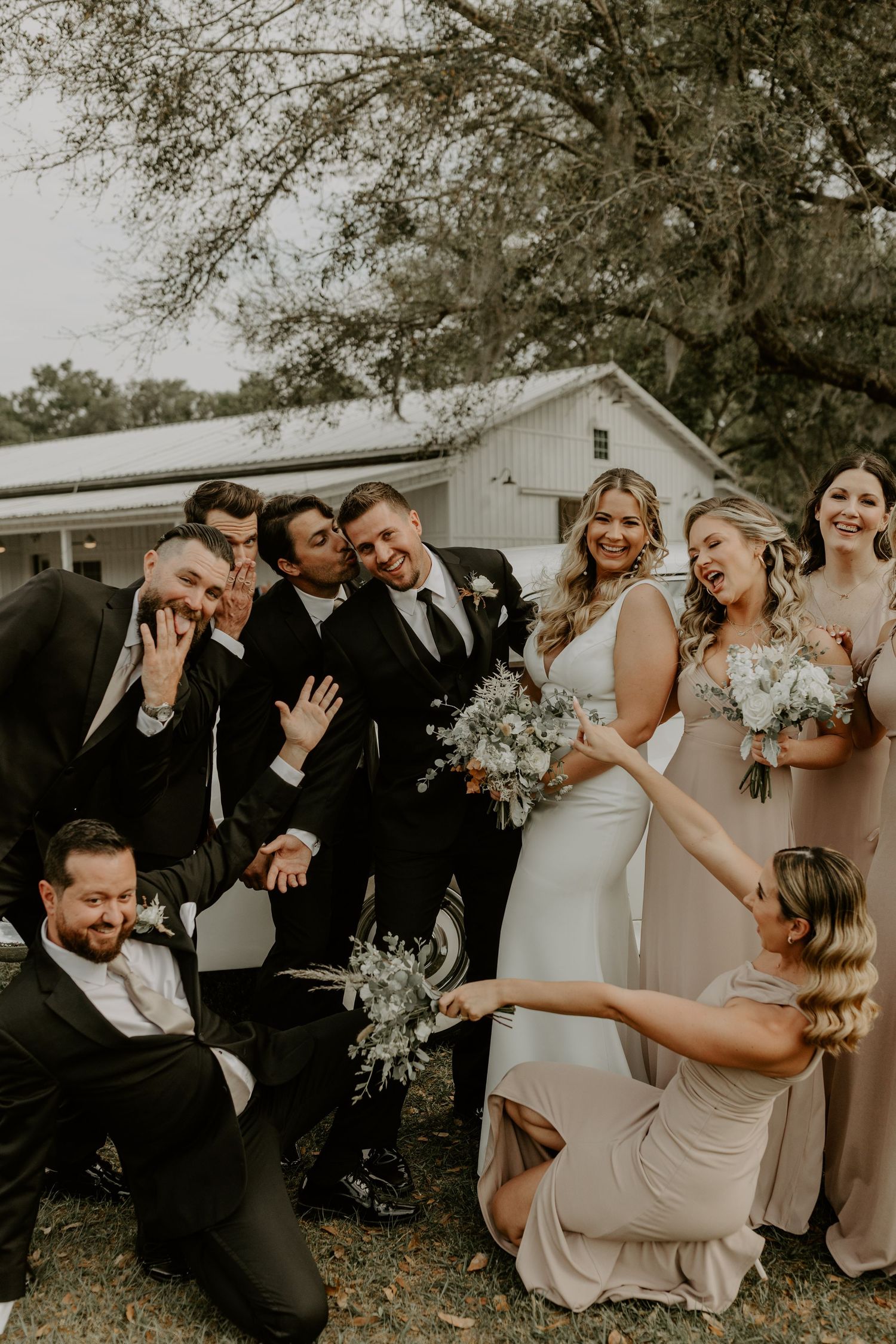 Wedding party poses candidly outdoors near white barn, showing joyful laughter and celebration in warm sepia tones.