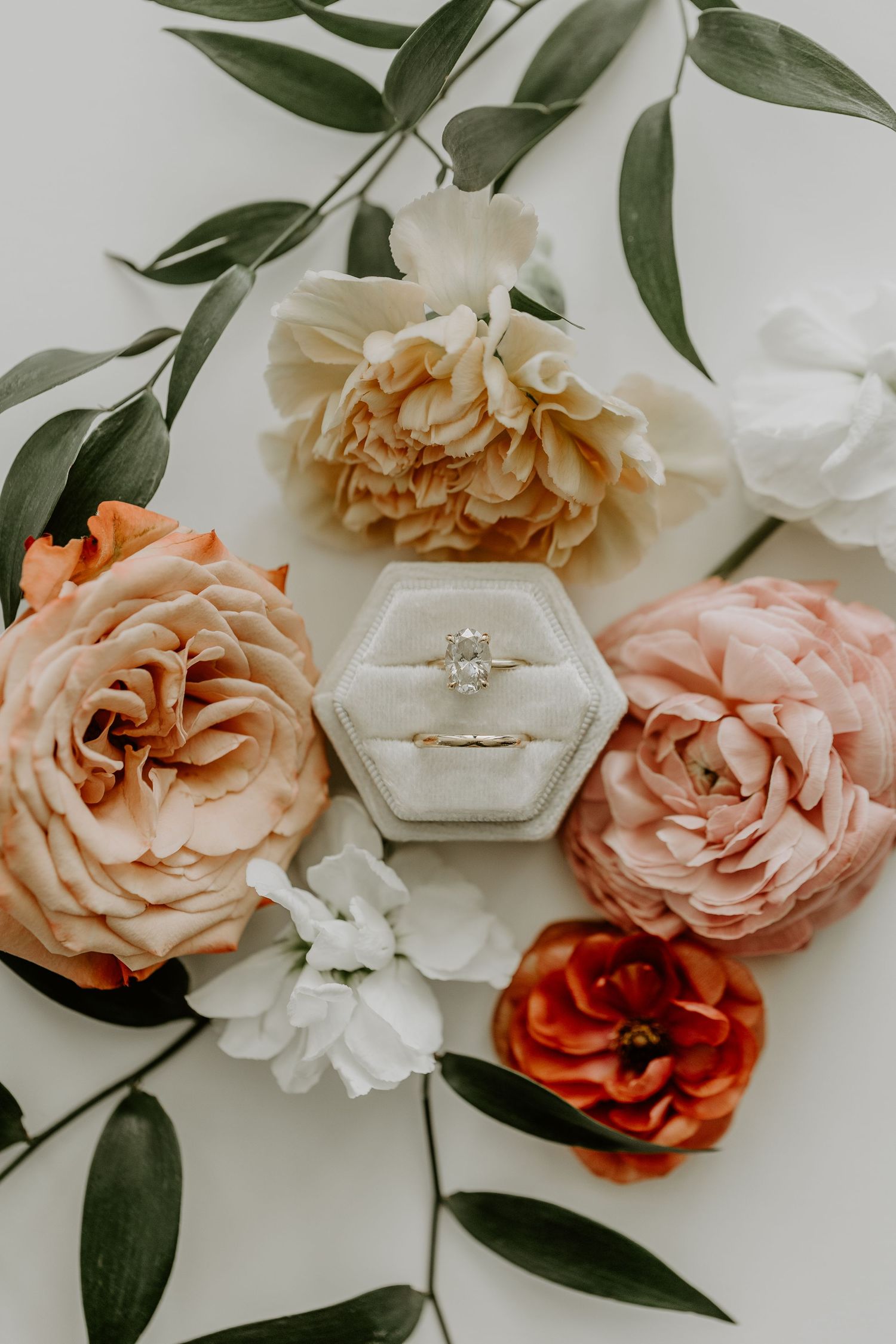 An engagement ring in hexagonal box surrounded by pink, peach and white roses with green foliage on white background.