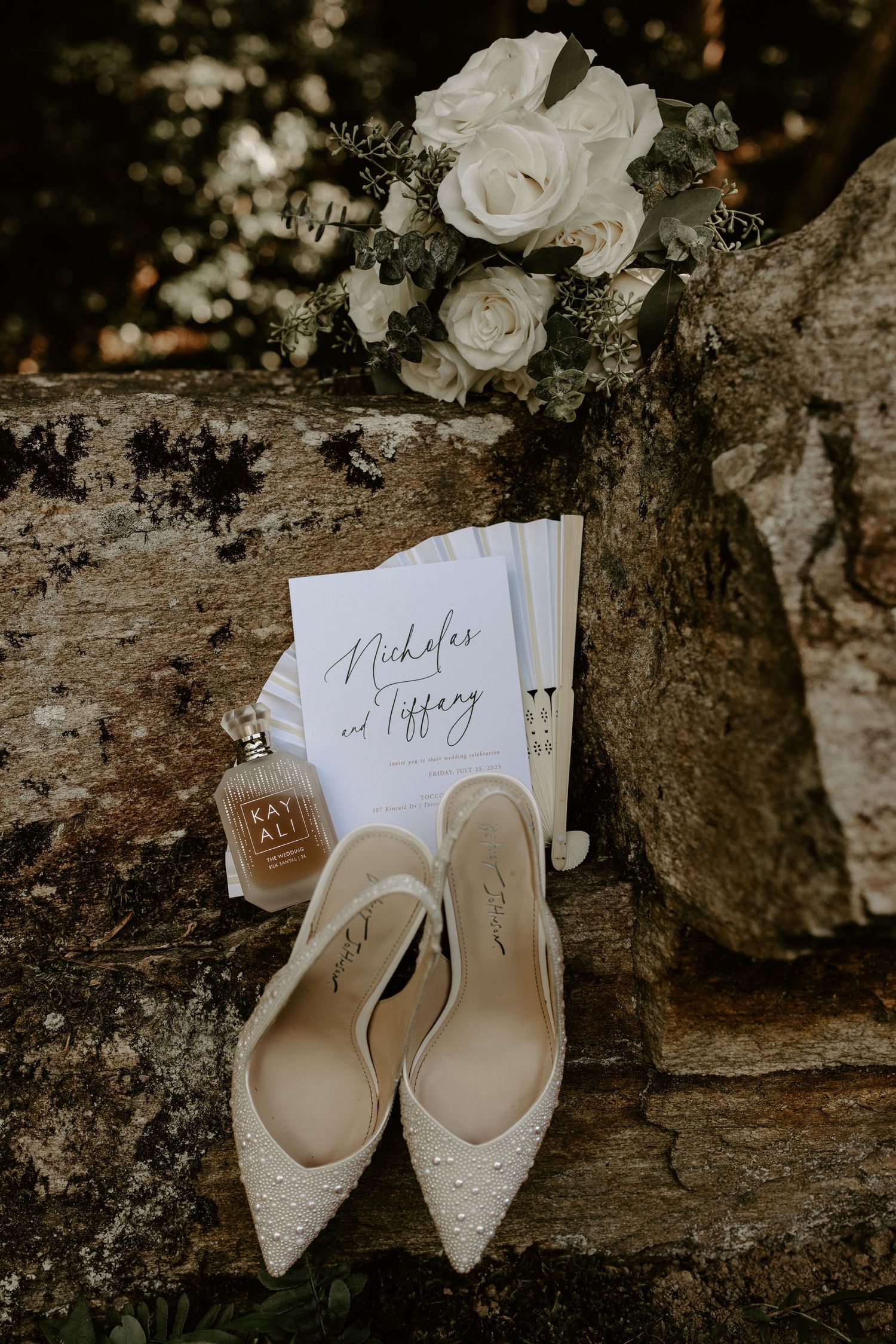 Wedding details featuring white roses bouquet, elegant bridal shoes and invitation card arranged on rustic stone surface.