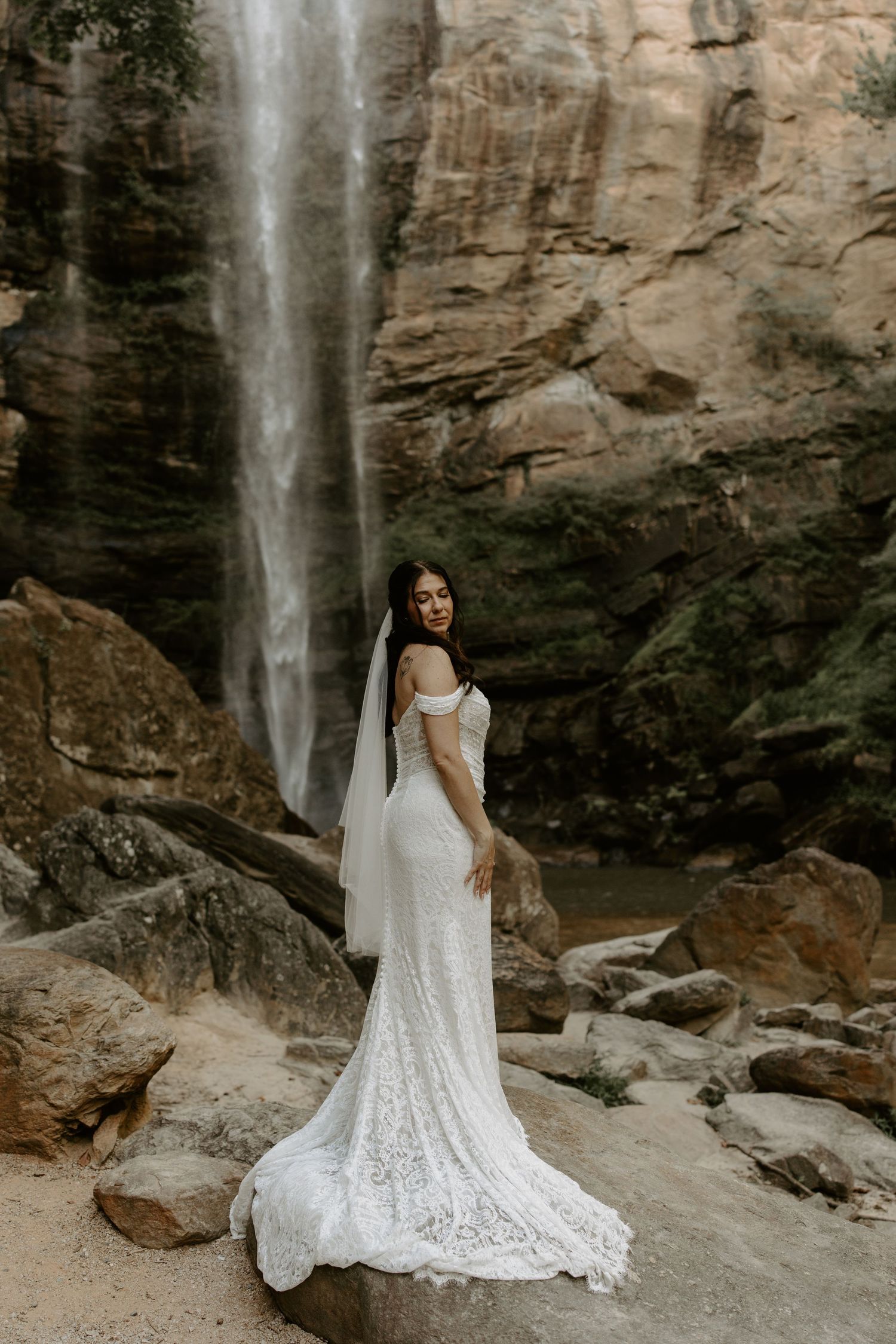 A woman in a white lace wedding dress poses against dramatic rocky cliffs and a cascading waterfall in a scenic outdoor setting.