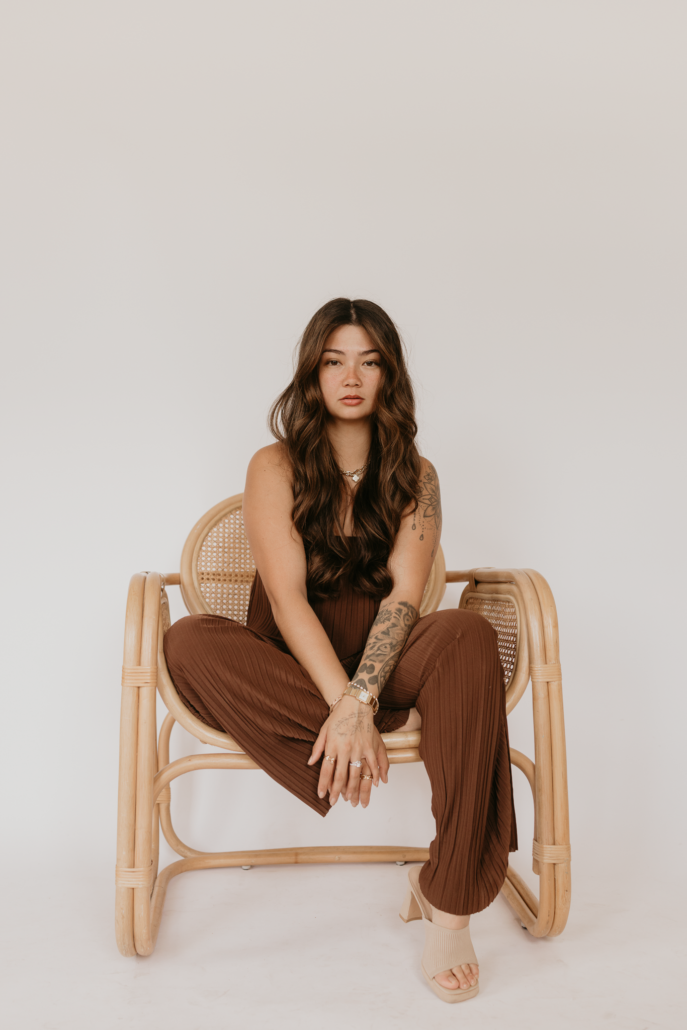 Woman in brown wide-leg pants and top sits in rattan chair, showcasing tattoos and wavy hair.