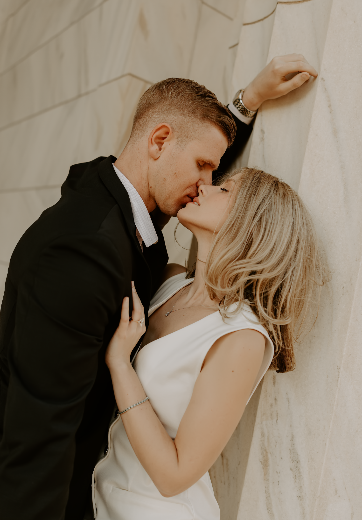 Couple in formal attire sharing intimate moment against white wall.