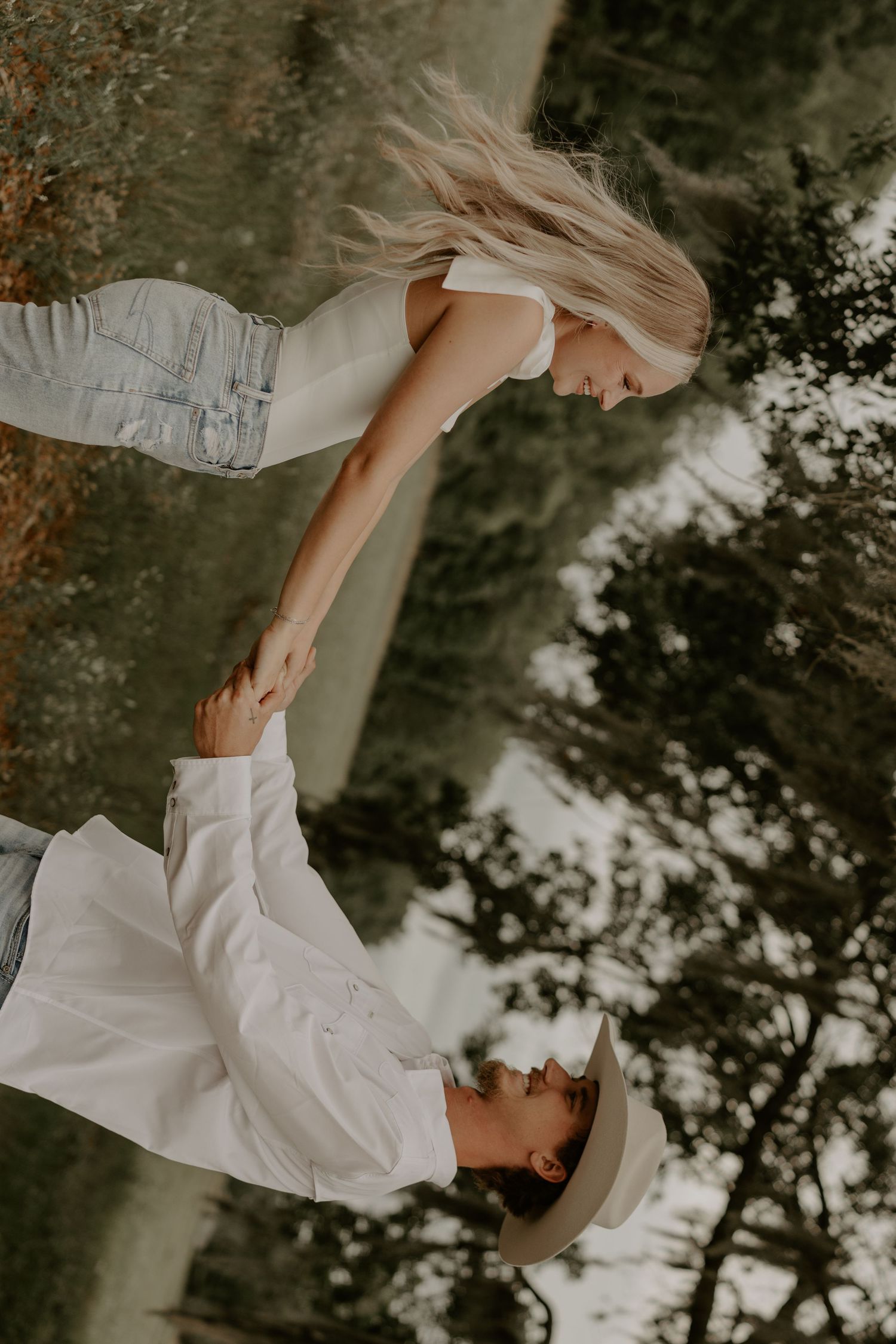 A romantic and ethereal scene of a couple in white clothing embracing outdoors against a backdrop of trees and sunlight.