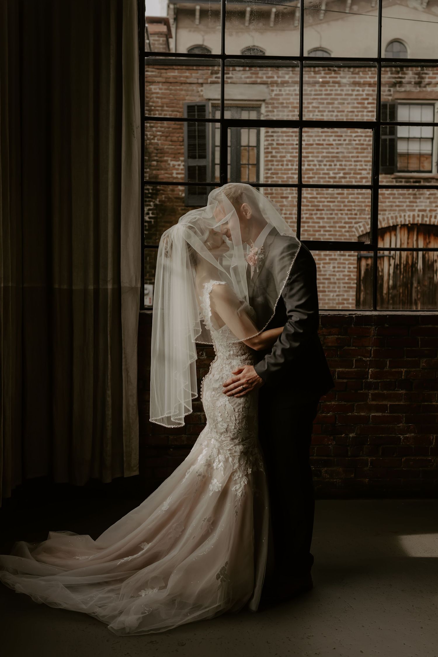 A bride and groom share an intimate kiss under her veil against a brick wall backdrop in moody lighting.
