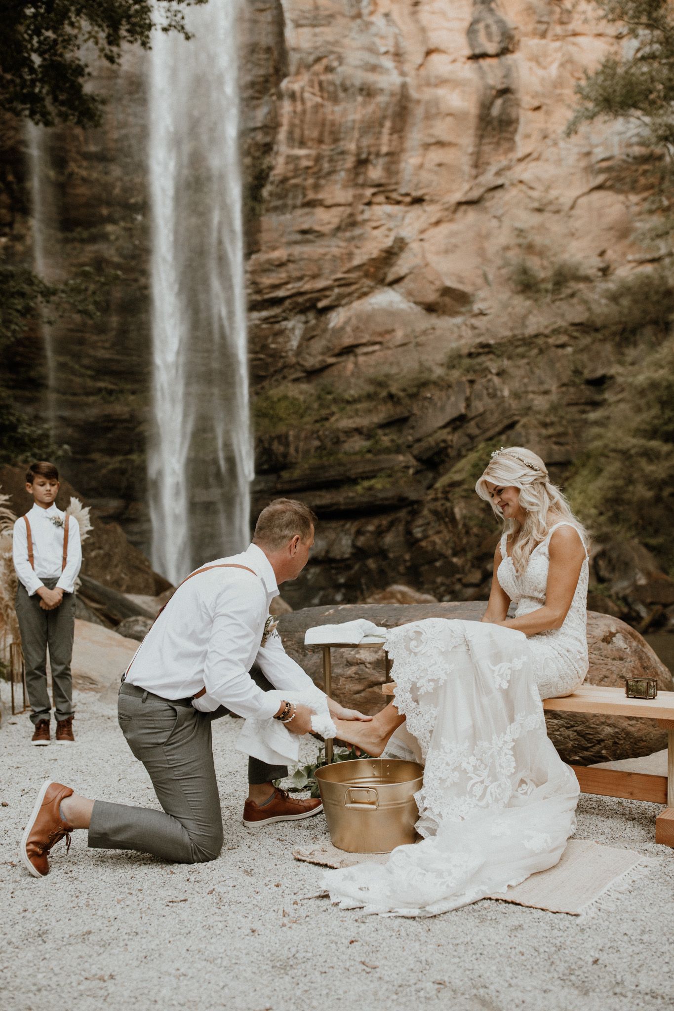A couple performs a ceremony near a majestic waterfall on a sandy clearing surrounded by tall rocky cliffs.