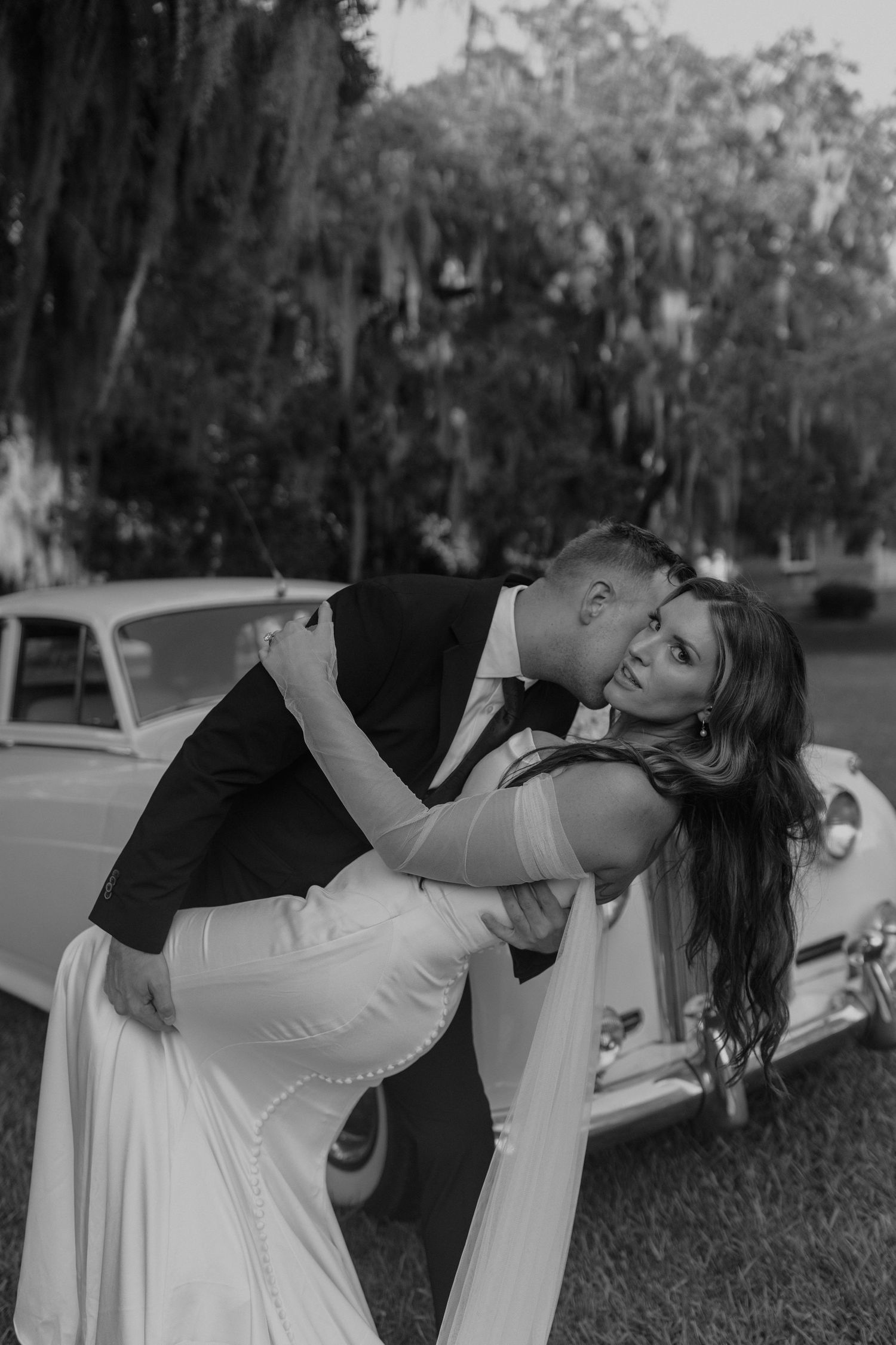 A romantic dip pose between couple in formal attire next to vintage car during outdoor wedding photoshoot.