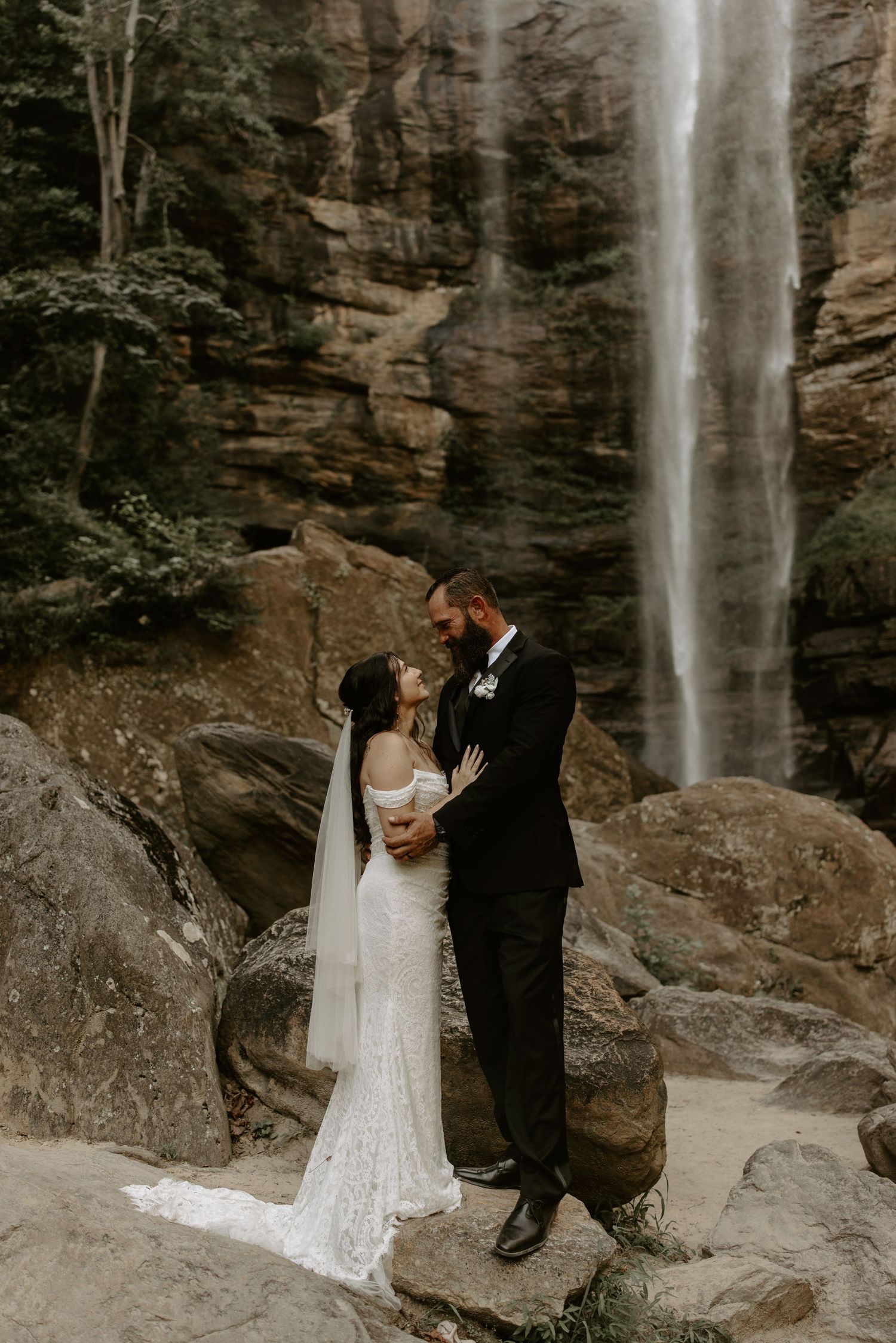 A romantic wedding moment captured at a scenic waterfall location with rocky cliffs in the background.